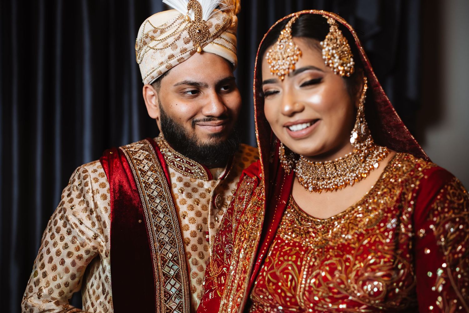South Asian bride and groom smiling closely in traditional red and gold wedding attire with jewelry.