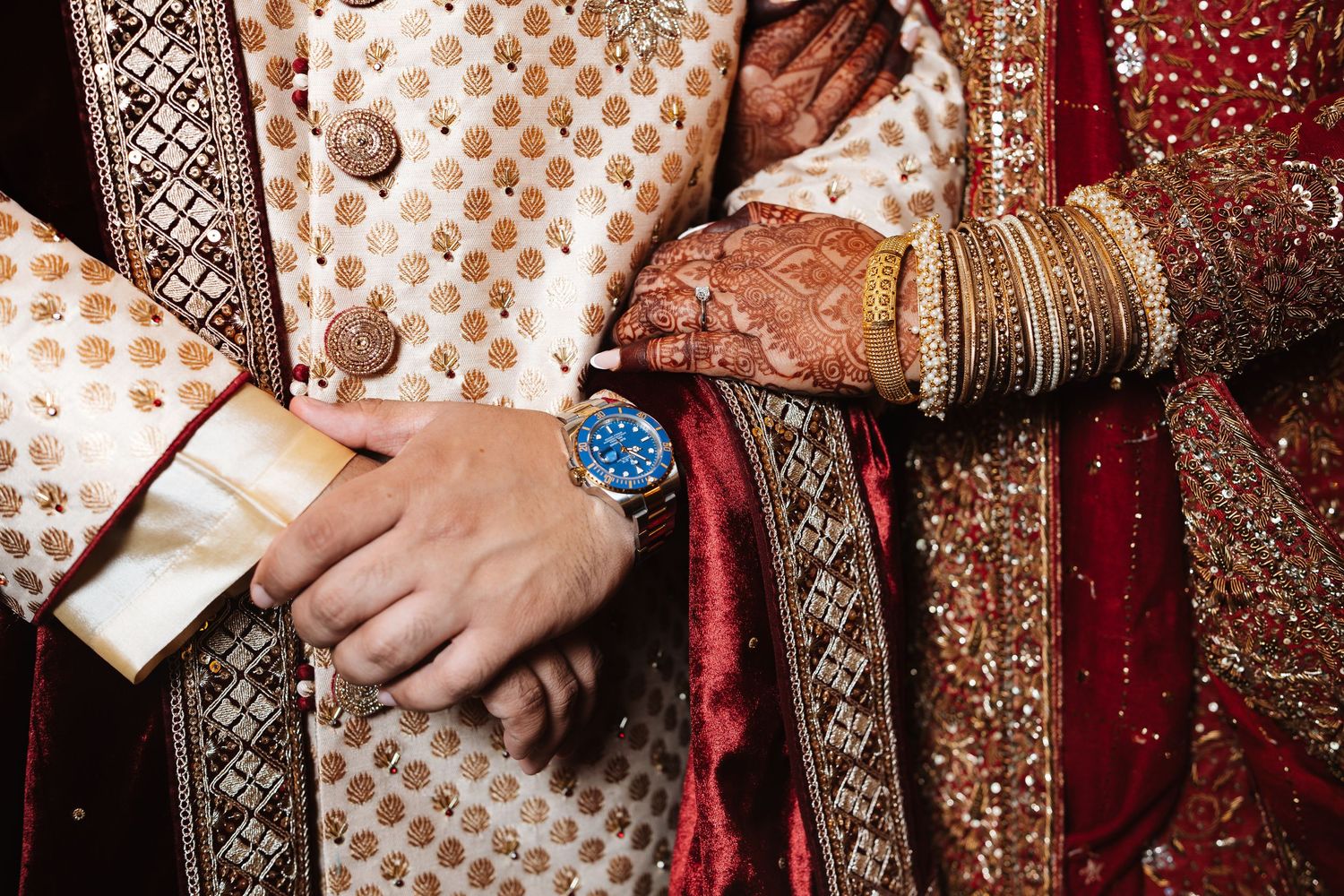 Detail shot of South Asian couple\u2019s hands showing mehndi, gold bangles, and groom\u2019s blue wristwatch.