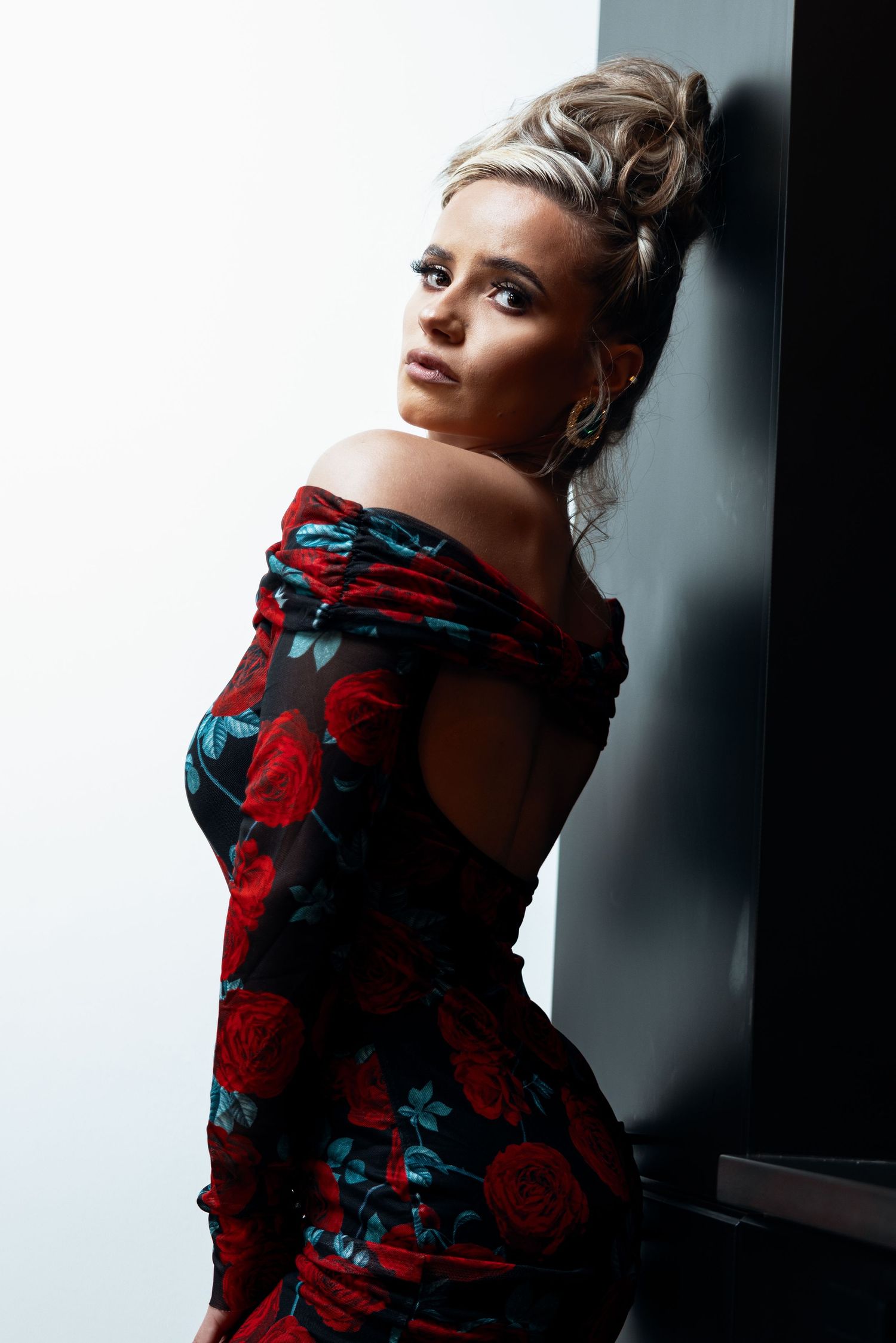 Blonde woman in off-shoulder red floral dress with updo leaning against dark wall.