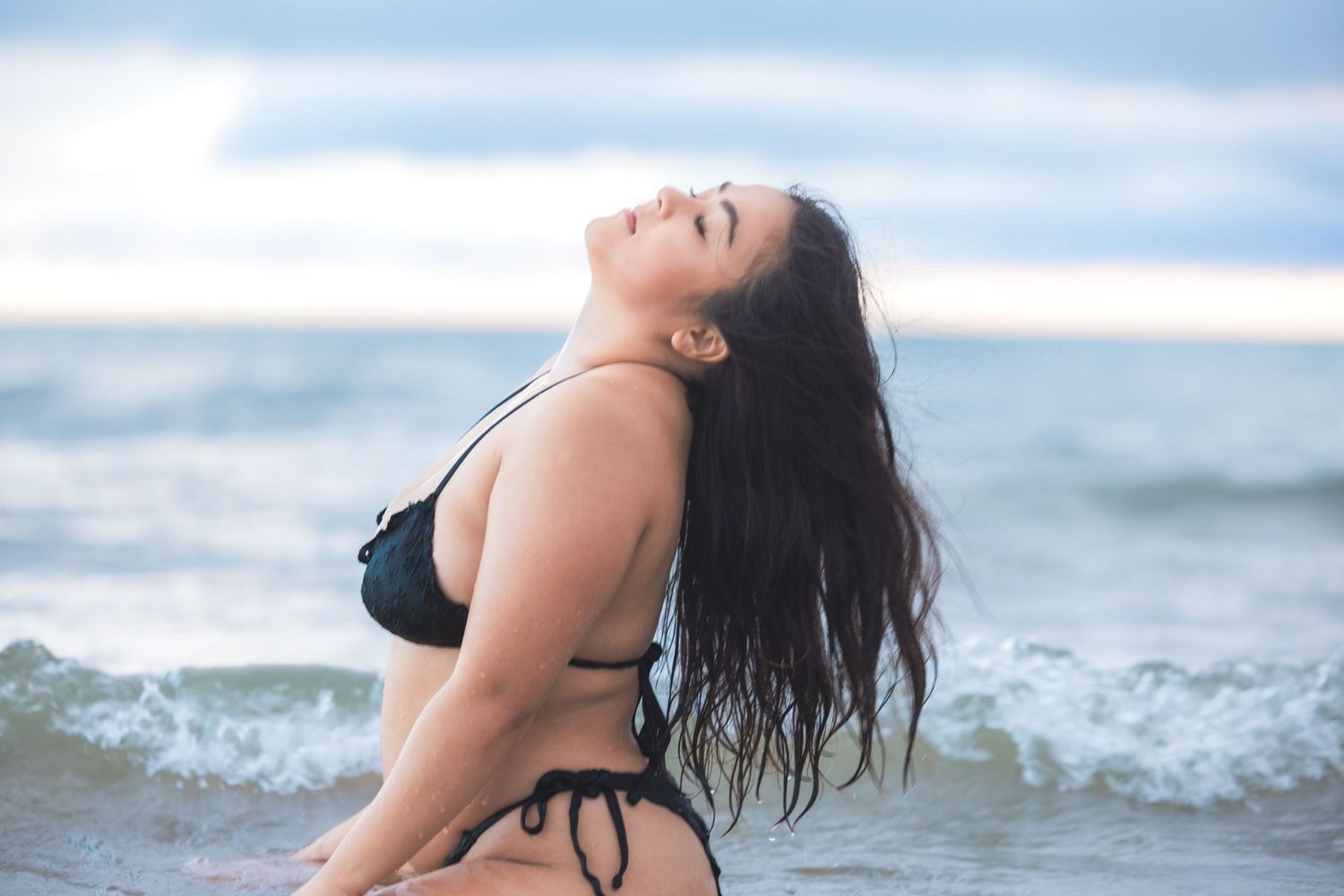 Plus size woman in black bikini with head tilted back standing in ocean surf on cloudy day.