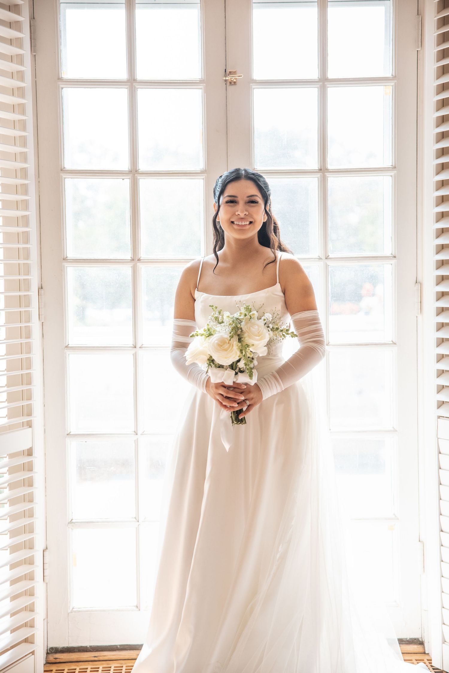 Smiling bride in white gown and sheer gloves holds white bouquet before bright windows.