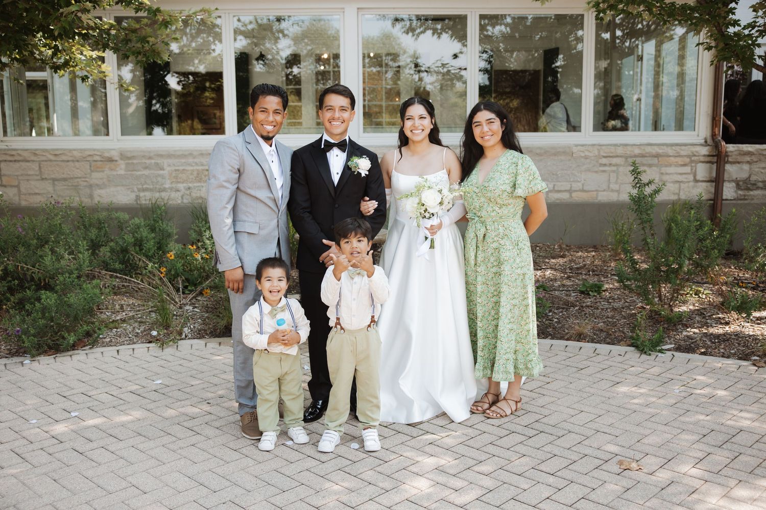 Bride and groom smile with family of four including two young boys outside wedding venue.