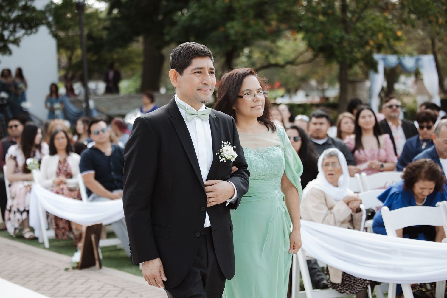 Groom in black tuxedo walks down aisle with mother in mint green gown at wedding.