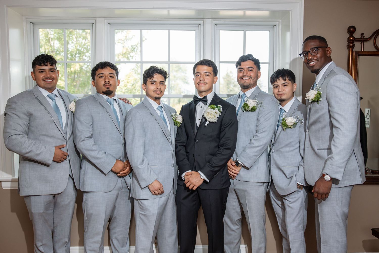 Groom in black tuxedo posing with six groomsmen in gray suits.