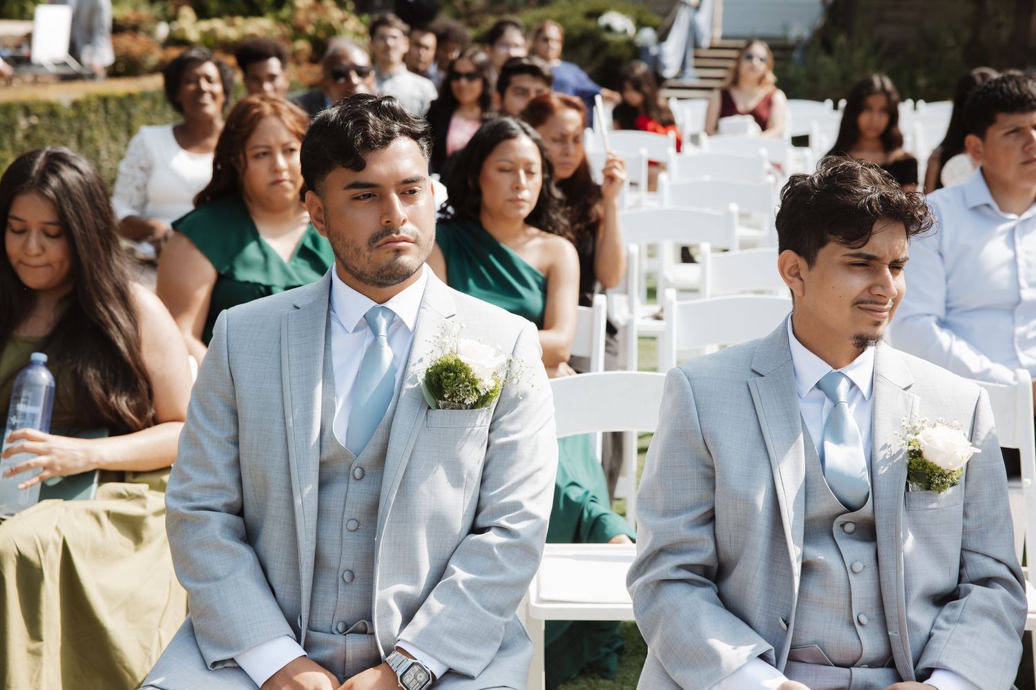 Two groomsmen in light gray suits with boutonnieres seated at outdoor wedding ceremony.