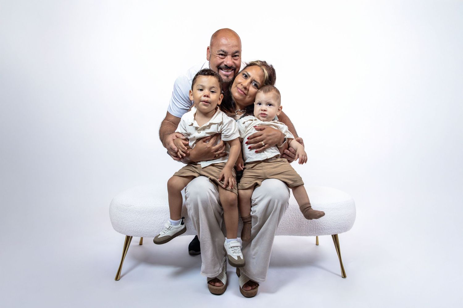 Smiling family of four sitting on white bench, parents holding two young children in matching beige outfits.