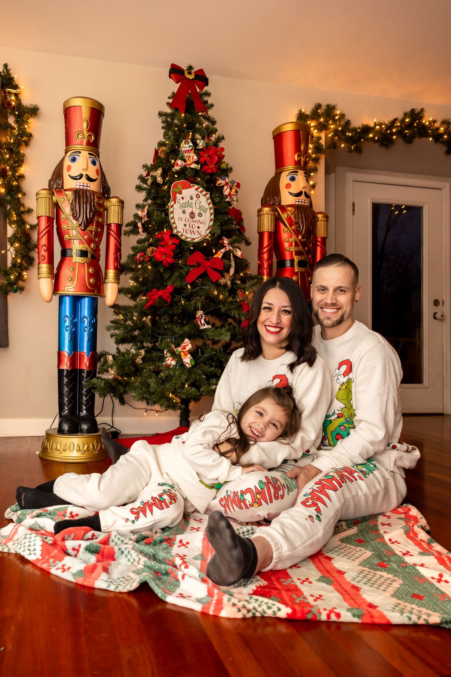 Smiling family of three in matching Christmas pajamas posing on a holiday blanket with nutcracker decorations.