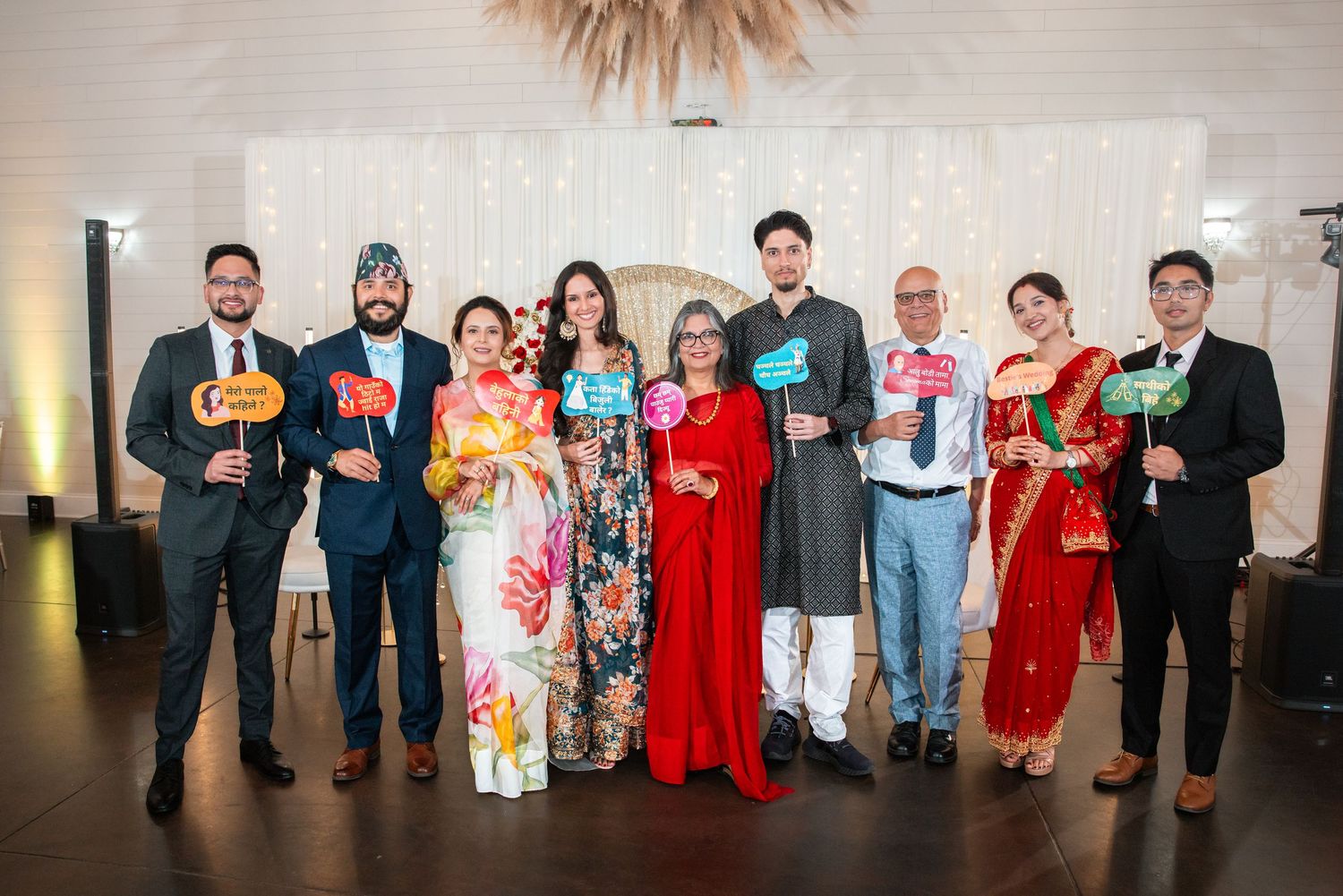 Diverse group of eight people in formal attire posing with colorful photo booth props at a wedding reception.