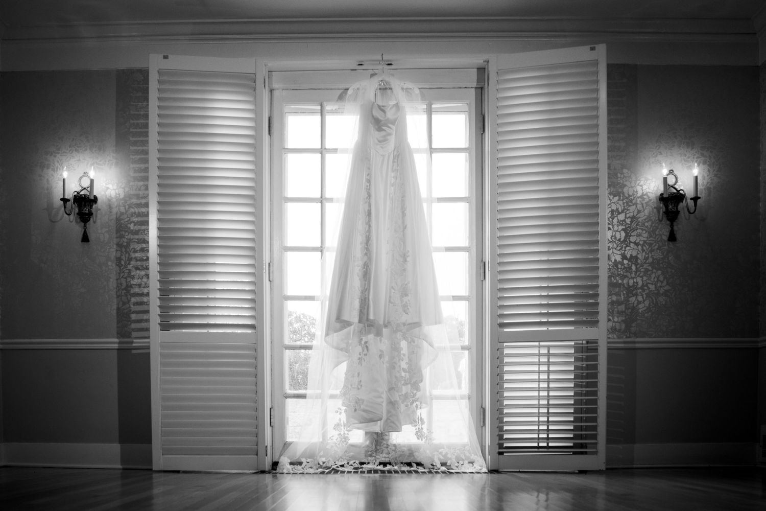 Black and white photo of a lace wedding gown hanging between shuttered French doors with wall sconces on each side.