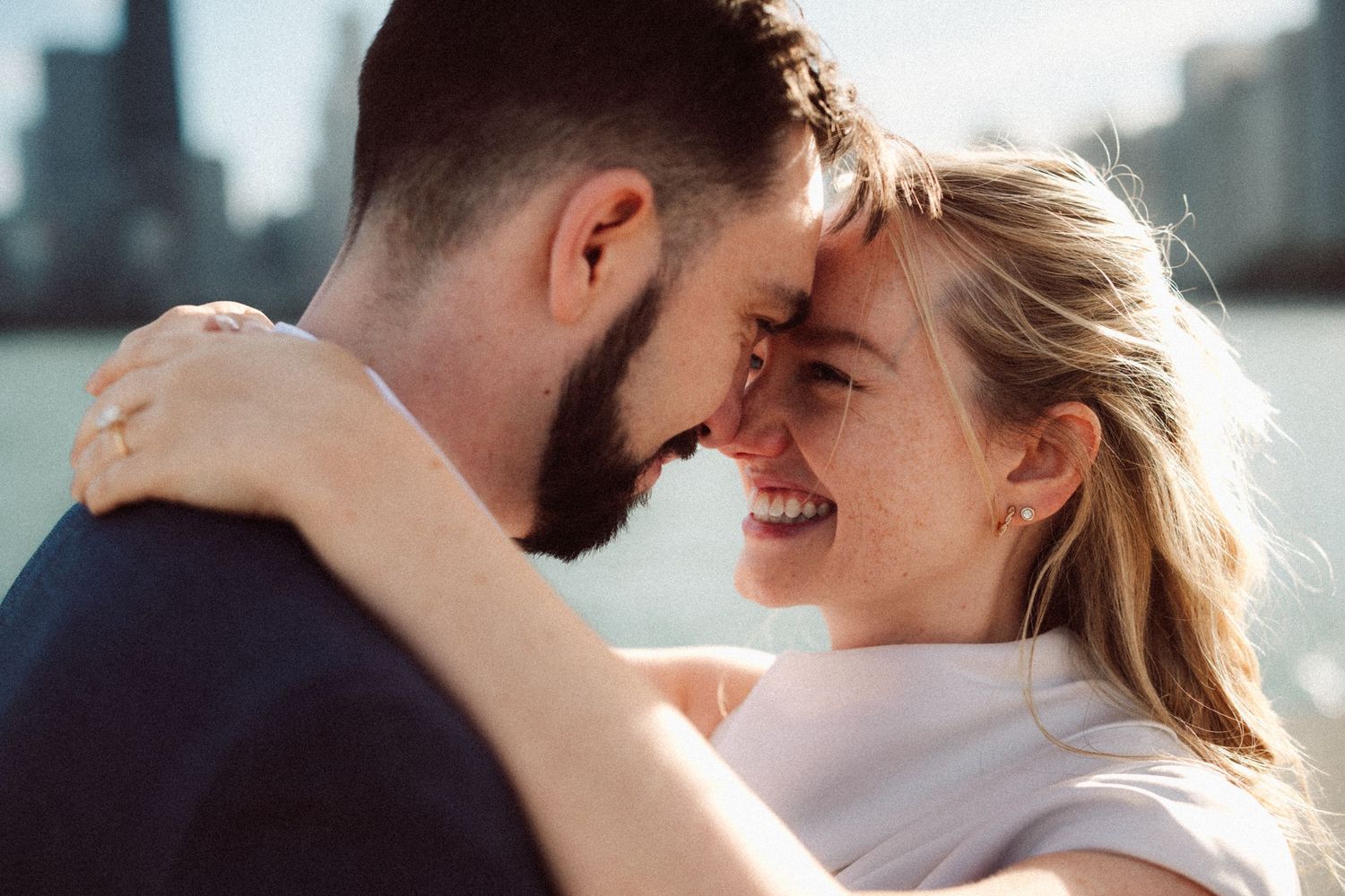 Happy couple sharing a joyful forehead-to-forehead moment by a city waterfront skyline.