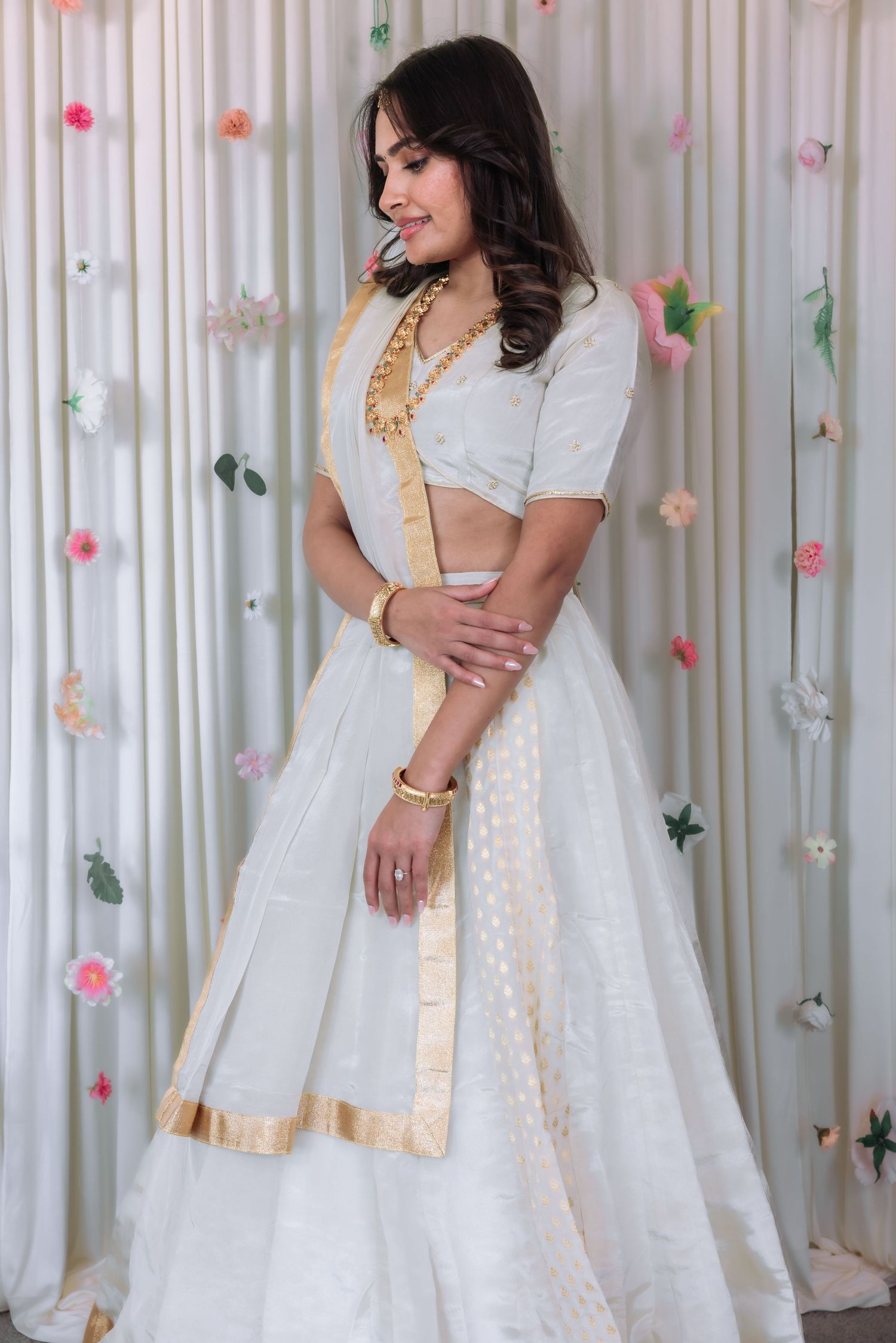Woman in elegant white and gold lehenga looking down gracefully before a floral curtain backdrop.