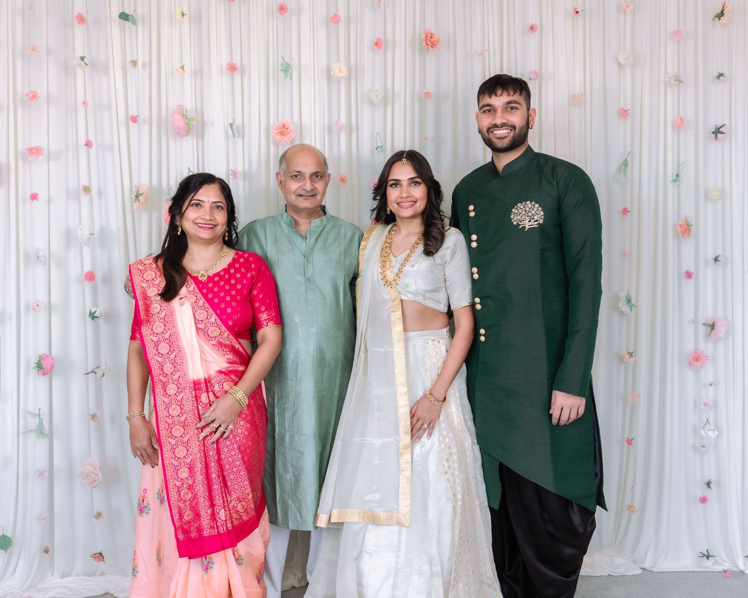 Family of four smiling together at Indian engagement ceremony with decorative white backdrop.