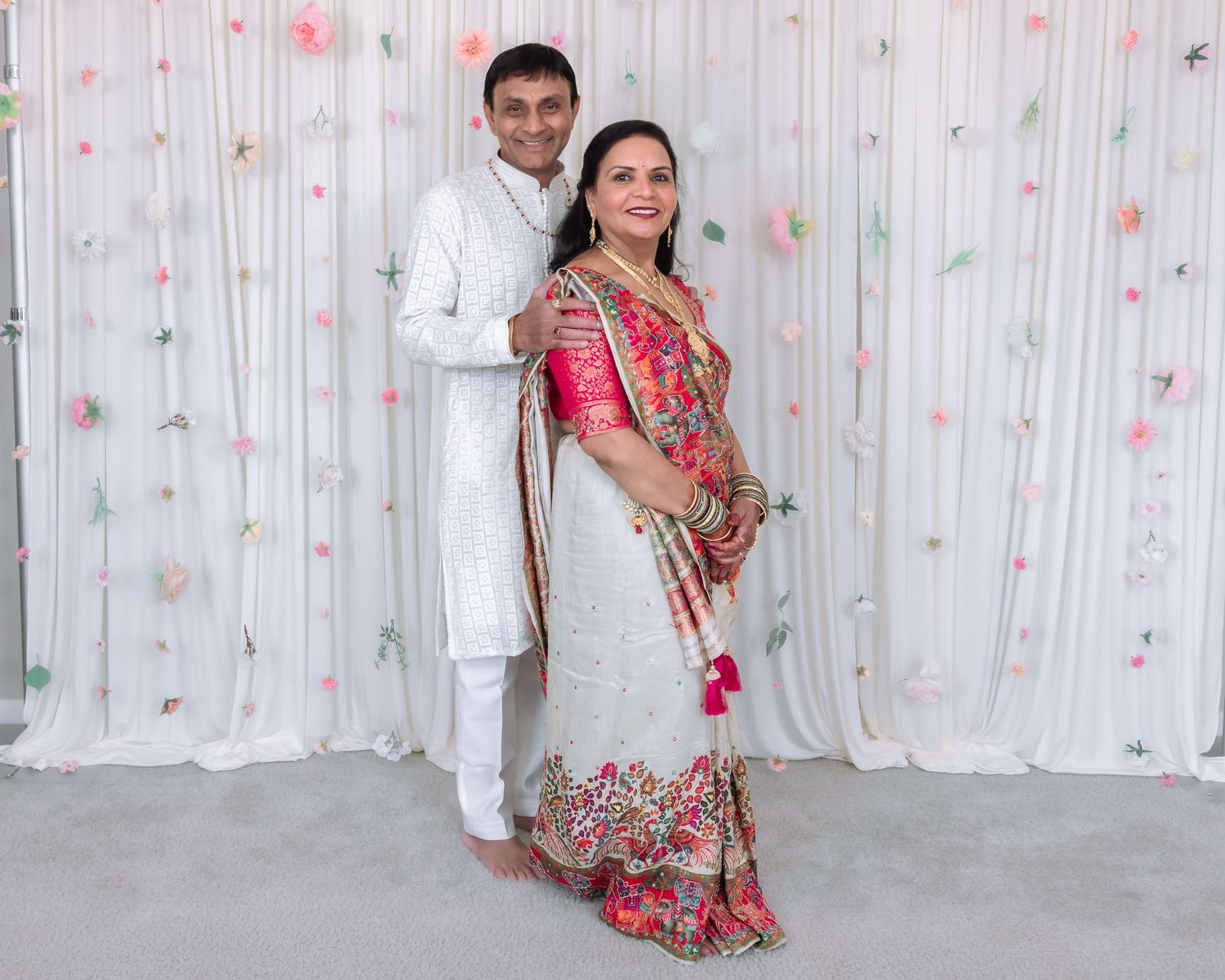 Indian couple posing together, man in white kurta and woman in elegant embroidered saree, floral backdrop.
