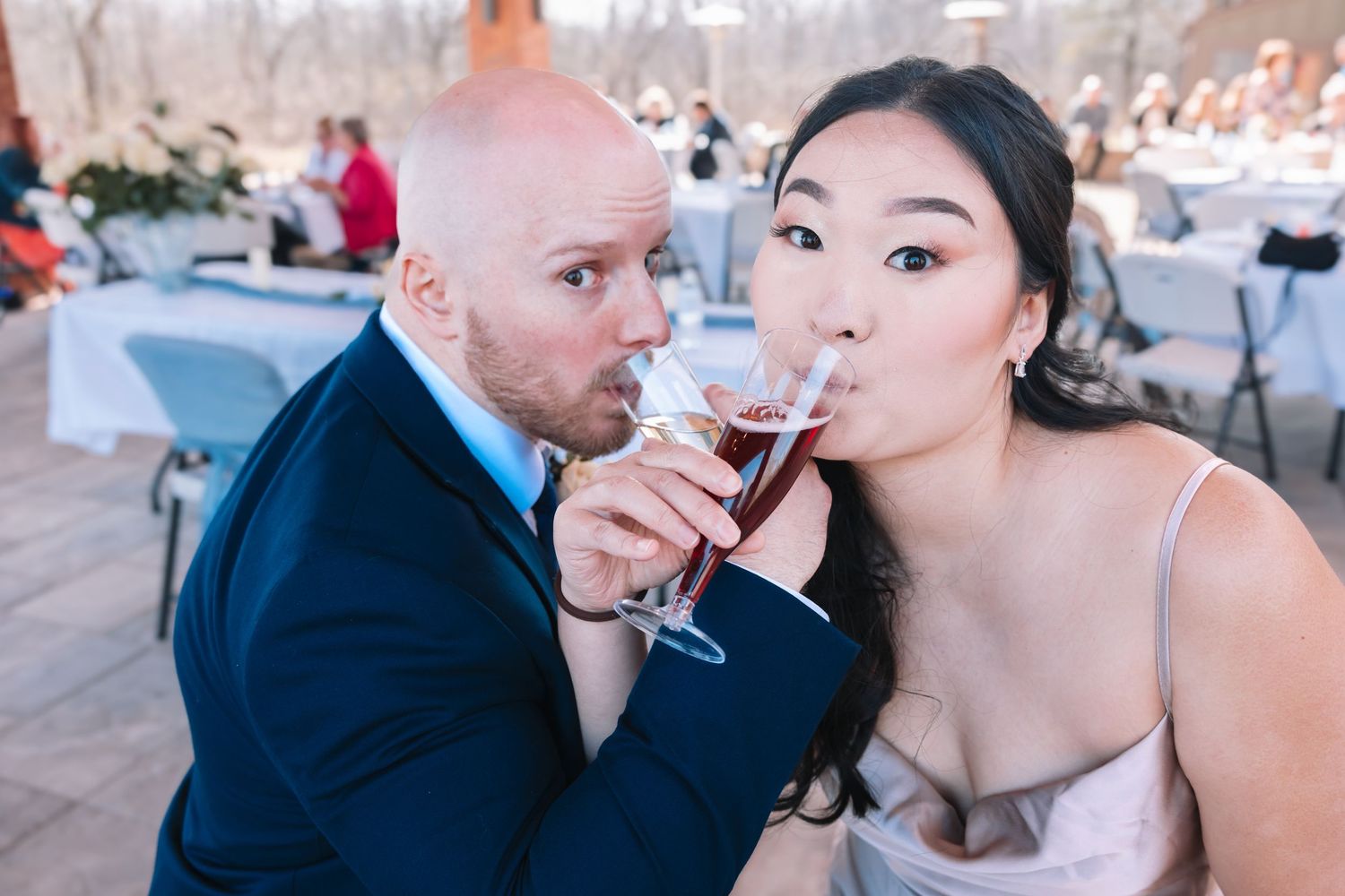 A bride and groom playfully share a drink from the same glass at their wedding reception.