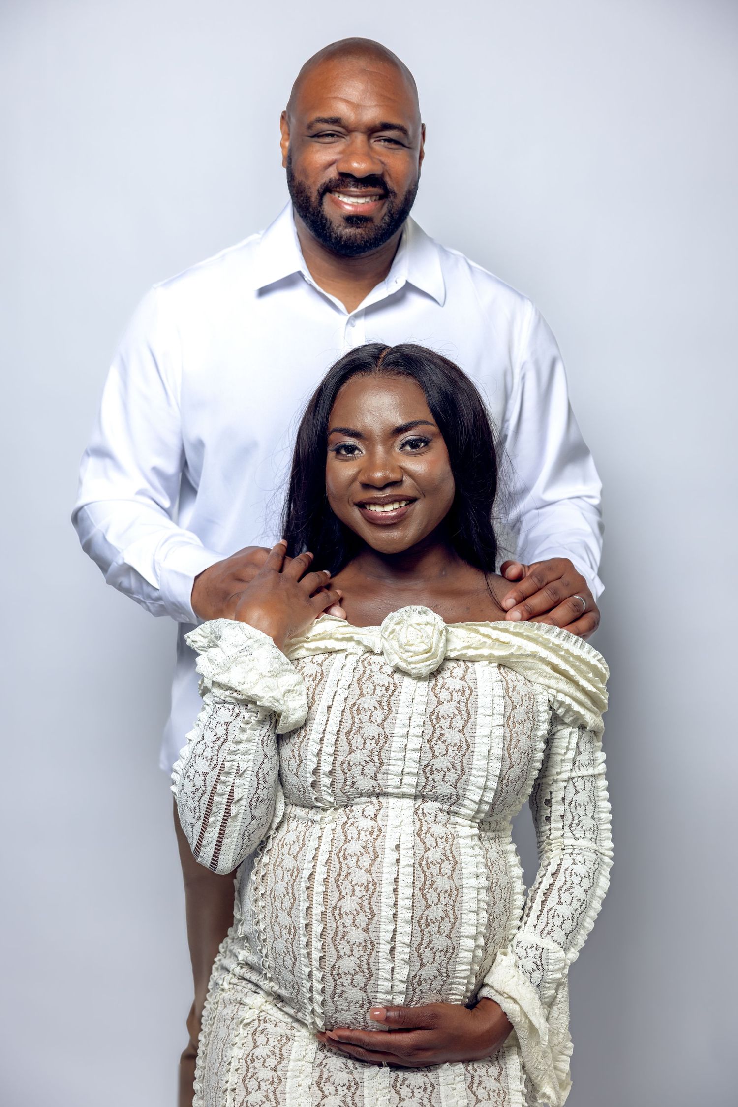 Smiling pregnant couple posing in studio, woman in white lace dress cradling baby bump, man behind her.