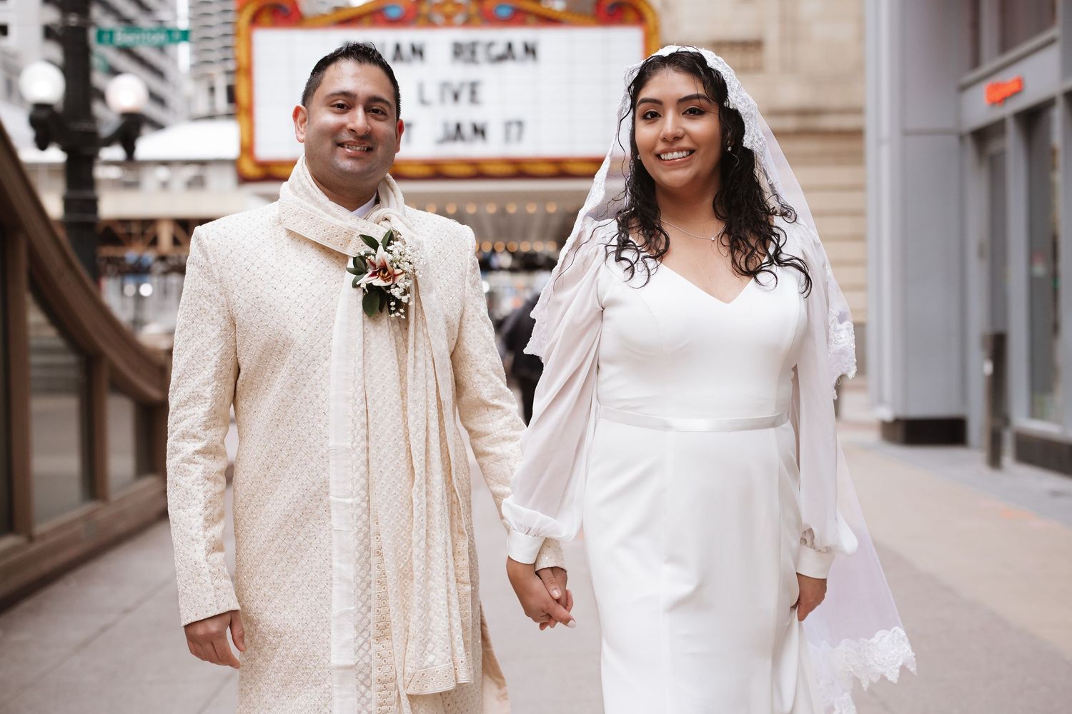 Indian wedding couple in white outfits hold hands while posing outside Chicago Theatre.