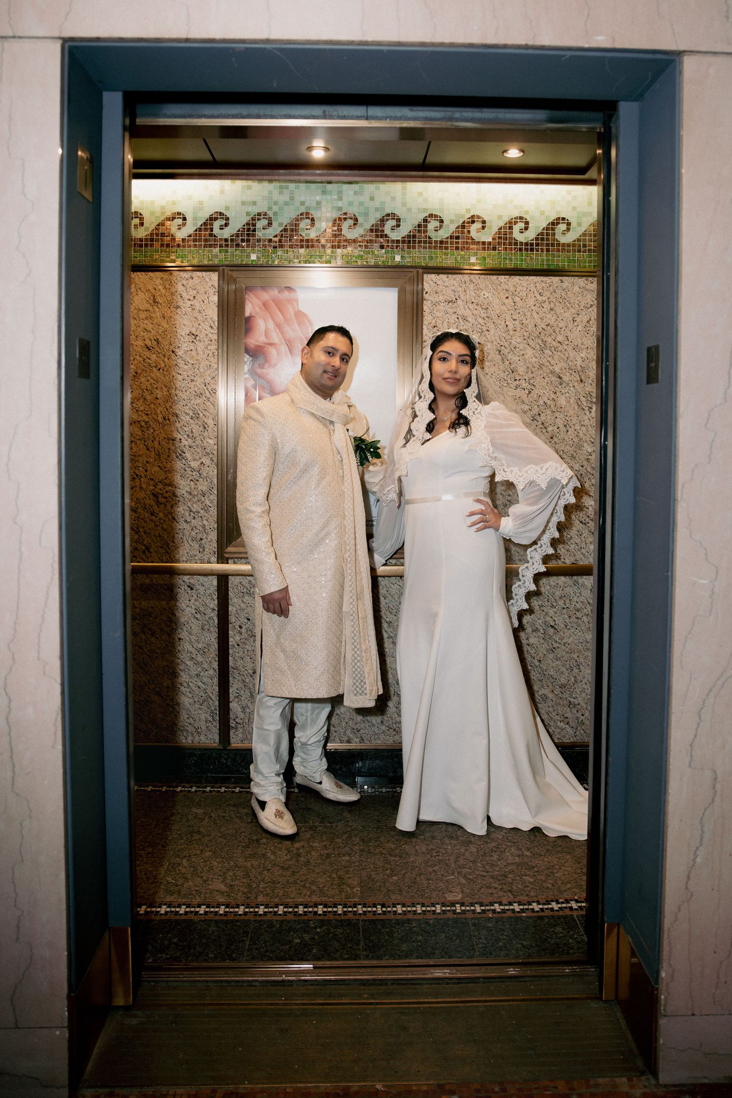 Bride in white gown with lace veil and groom in cream sherwani pose in vintage elevator.