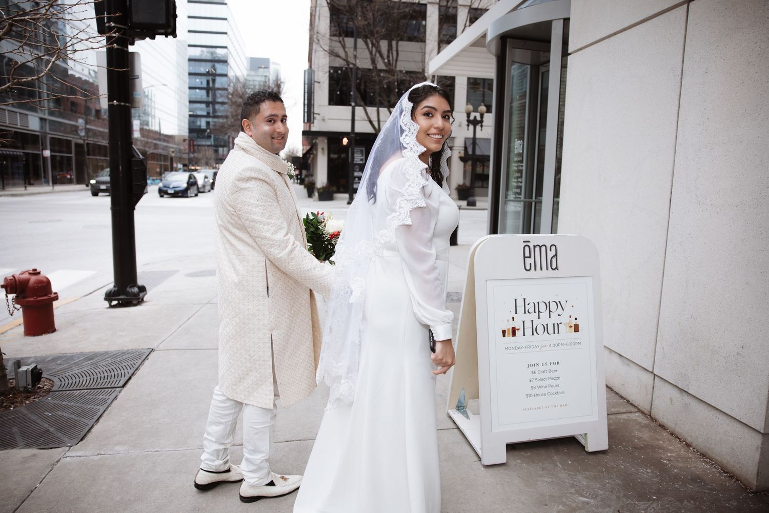 Bride in white gown with lace veil and groom in ivory suit posing outside near a restaurant happy hour sign.