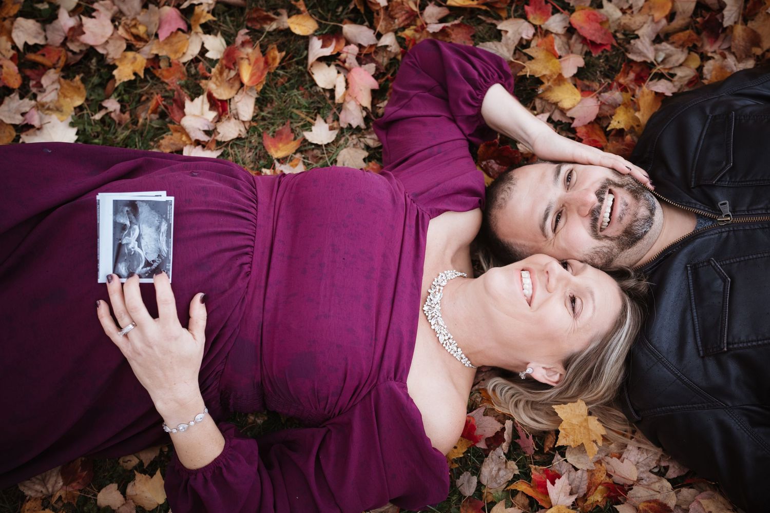 Couple lying in colorful autumn leaves, pregnant woman in purple dress holding ultrasound photo, smiling.