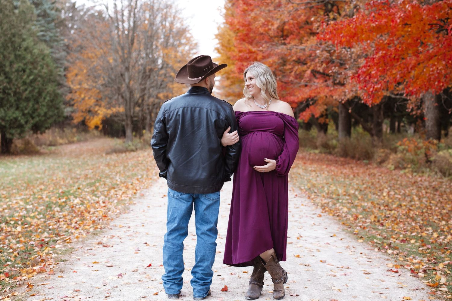 Pregnant couple walking away on a leaf-covered path surrounded by vibrant red autumn trees.