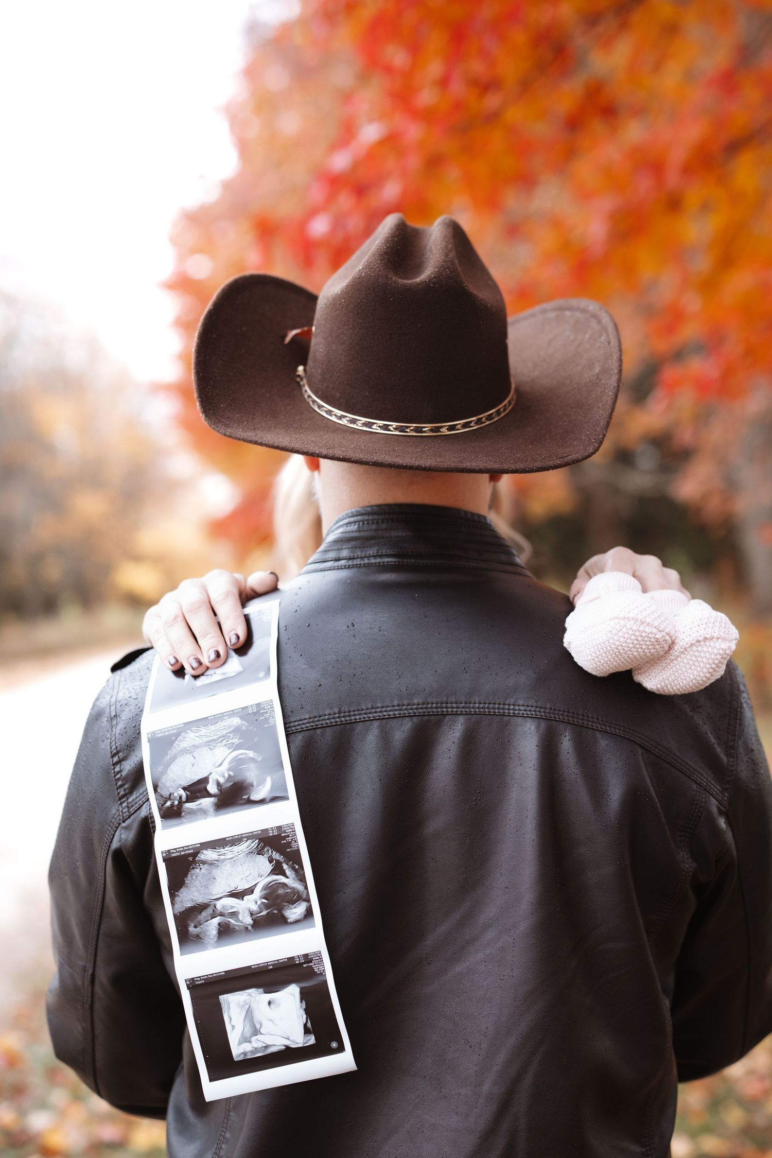 Man in cowboy hat and leather jacket holds ultrasound photos with baby shoes on his shoulder.