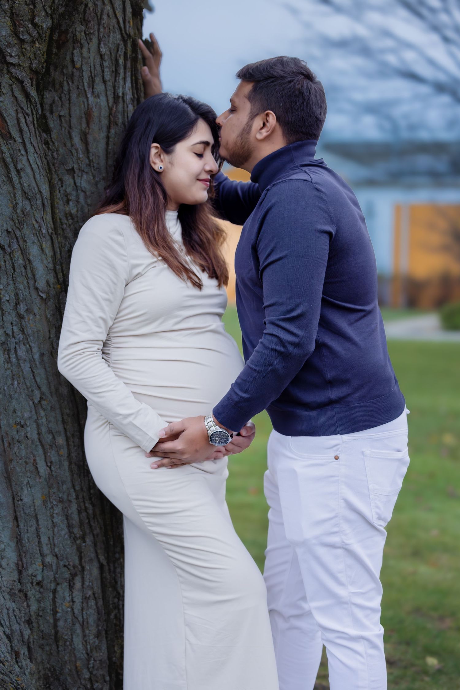 Man tenderly kissing pregnant woman leaning against a tree in a park.
