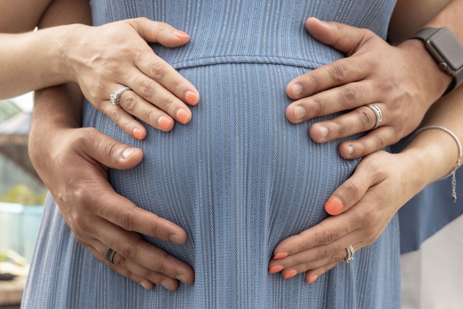 Close-up of couple's hands with wedding rings tenderly holding a pregnant belly in a blue maternity dress.