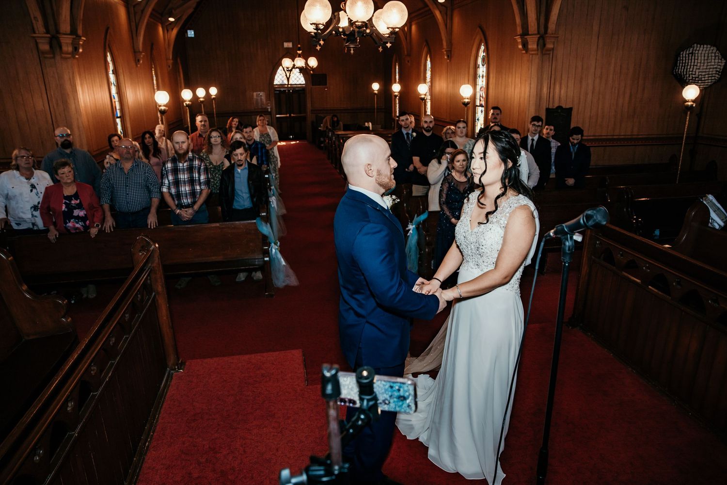 Overhead view of bride and groom holding hands at church altar with wedding guests standing in pews.