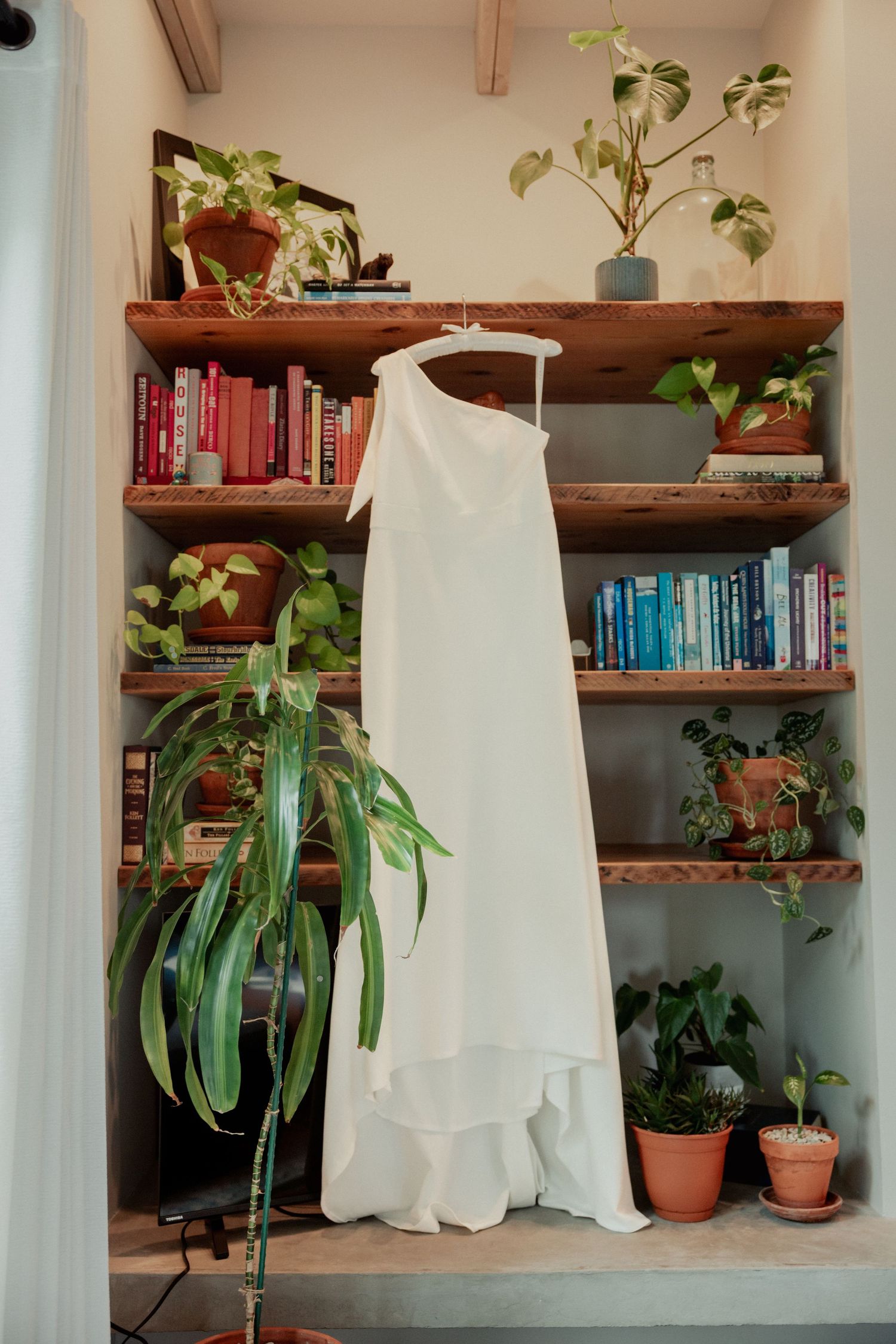 Minimalist white one-shoulder bridal gown displayed on a hanger in a cozy bookshelf nook.