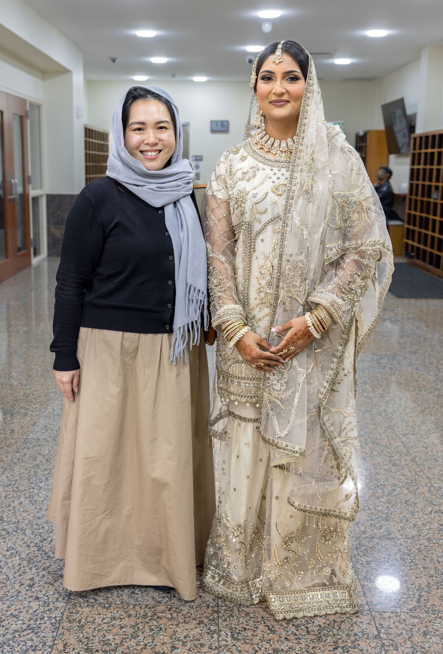 A bride in ornate white South Asian bridal attire poses with a woman in hijab and casual black top in a hallway.