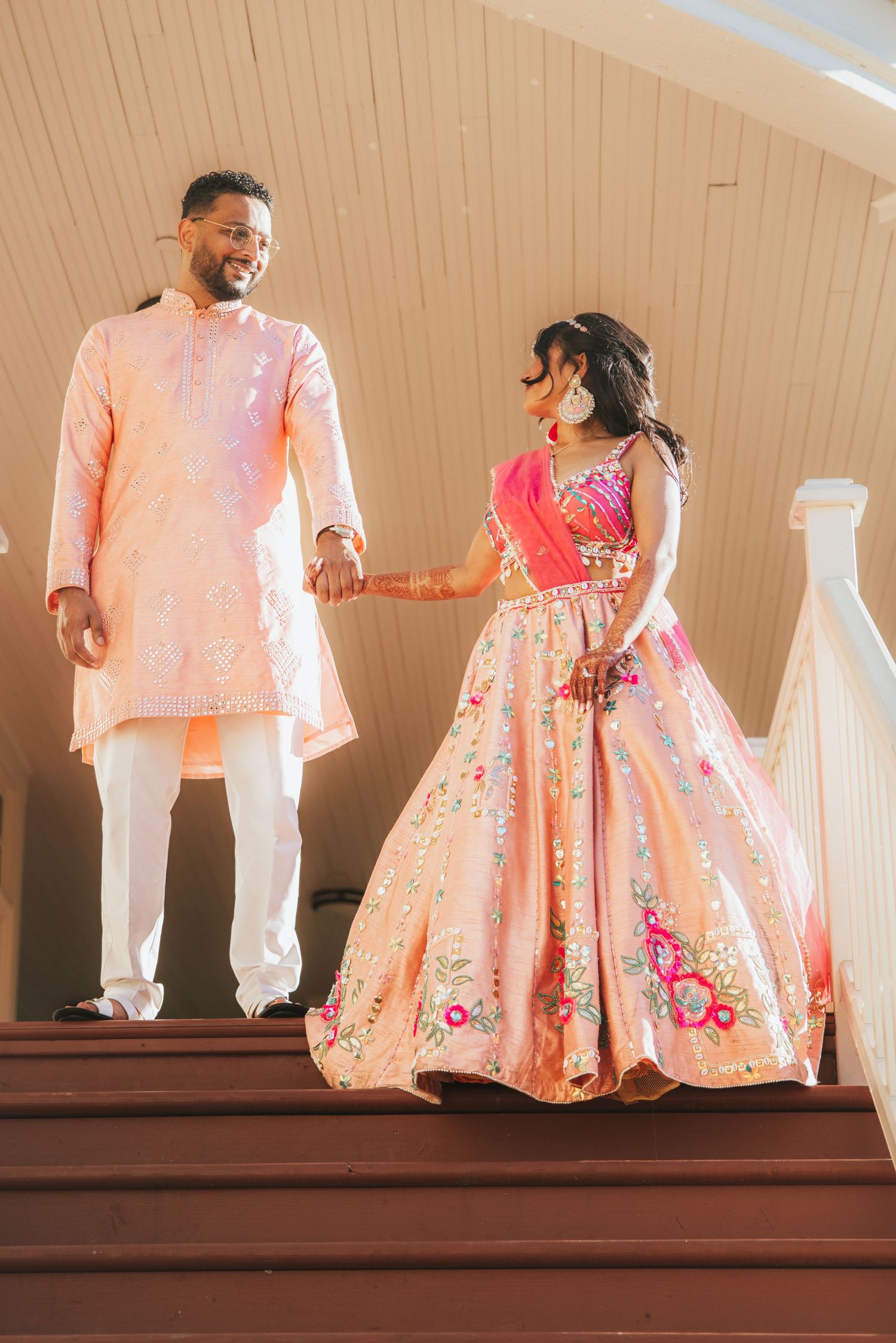 Indian couple in traditional peach outfits hold hands descending a staircase, brides lehenga flowing gracefully.