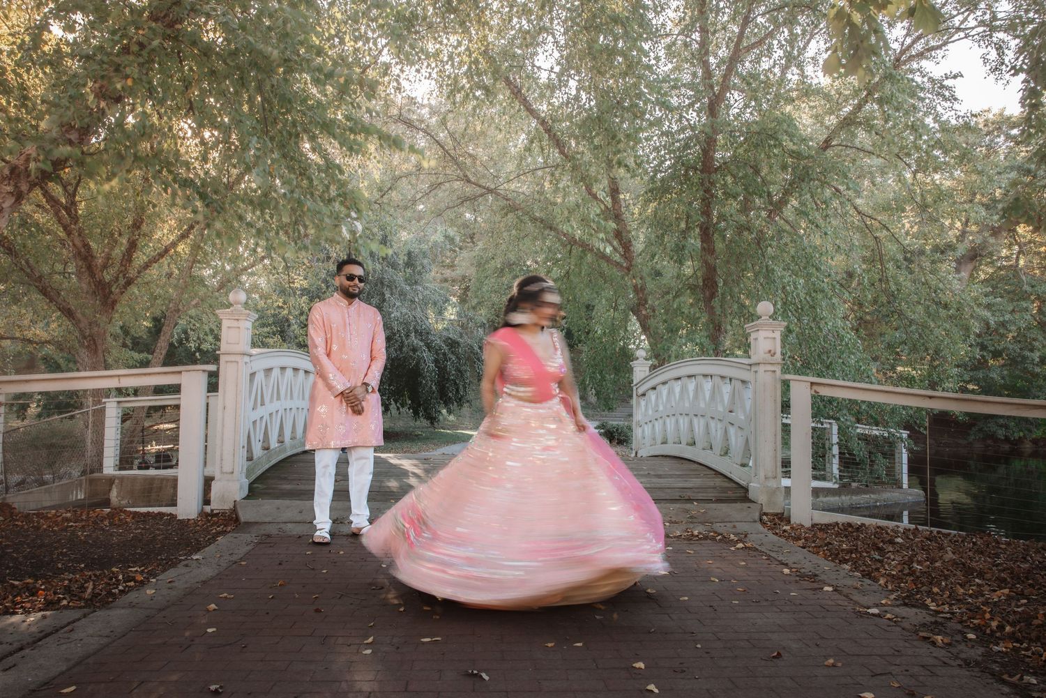 Bride twirling pink floral lehenga while groom watches near white decorative garden bridge.