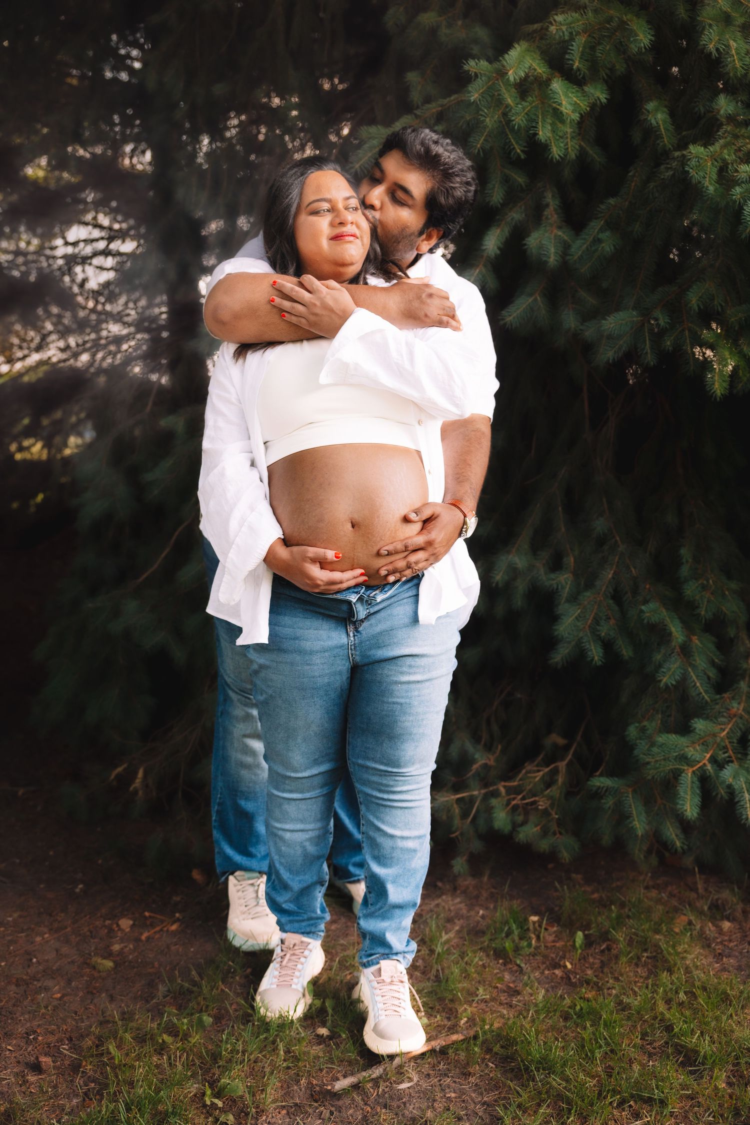 Couple in white outfits embracing outdoors, man hugging pregnant woman from behind near evergreen trees.