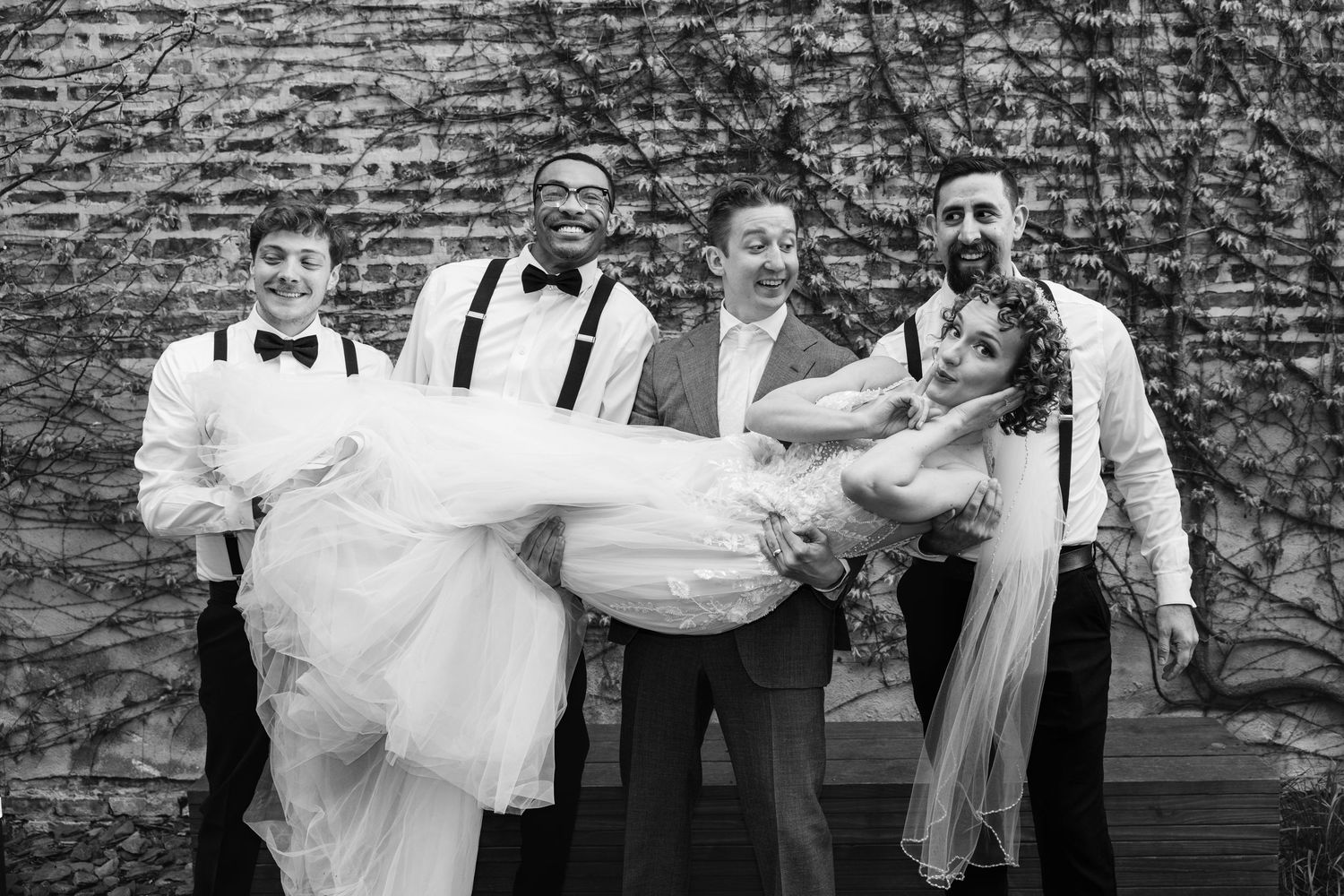 Black and white photo of groomsmen lifting smiling bride in wedding dress outdoors against brick wall.