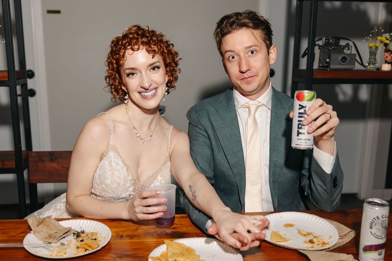 Smiling newlyweds sit together at a wooden table enjoying food and drinks during their wedding reception.