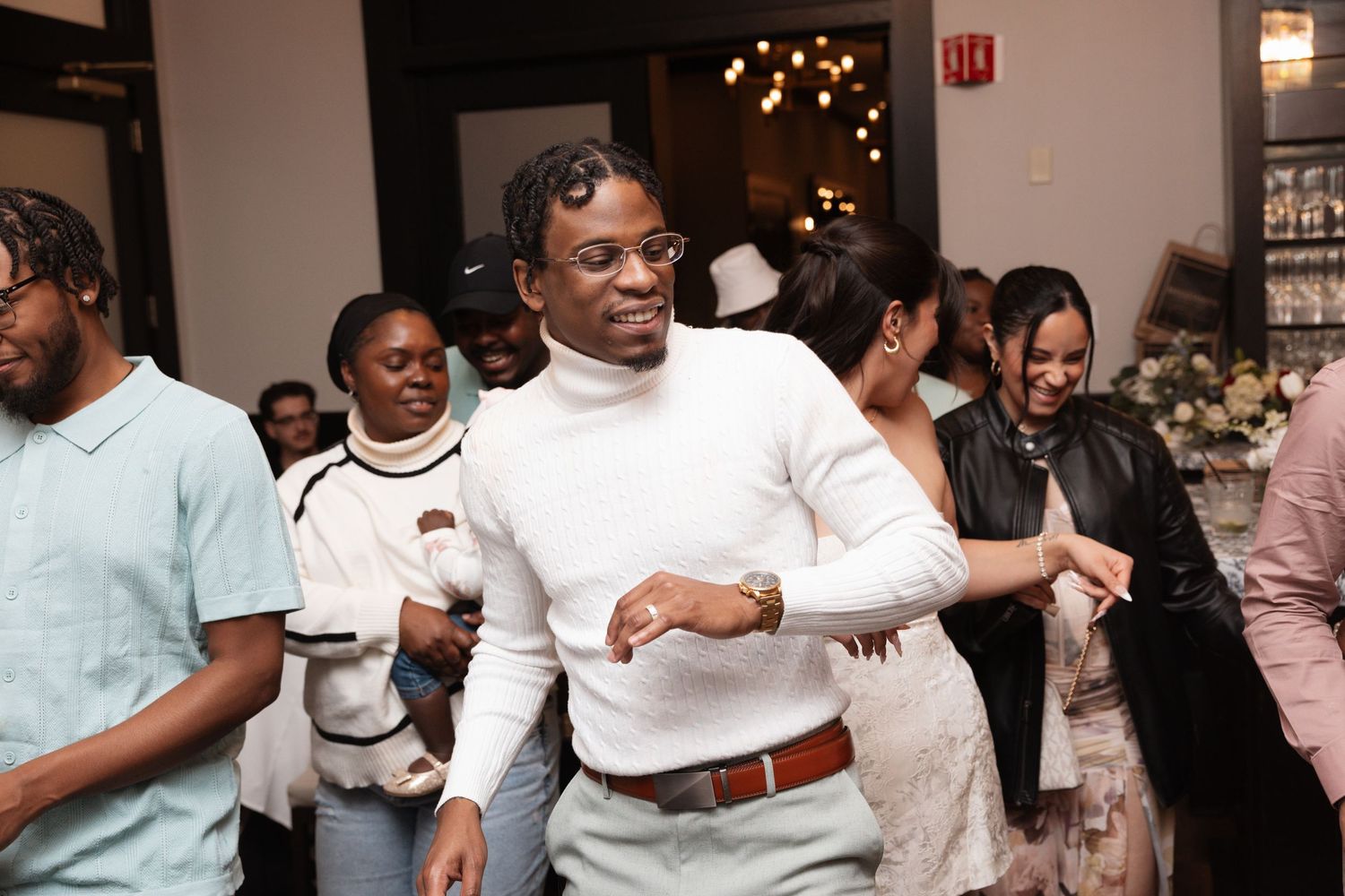 Groom in white turtleneck and gray pants dances joyfully with guests at lively wedding reception.