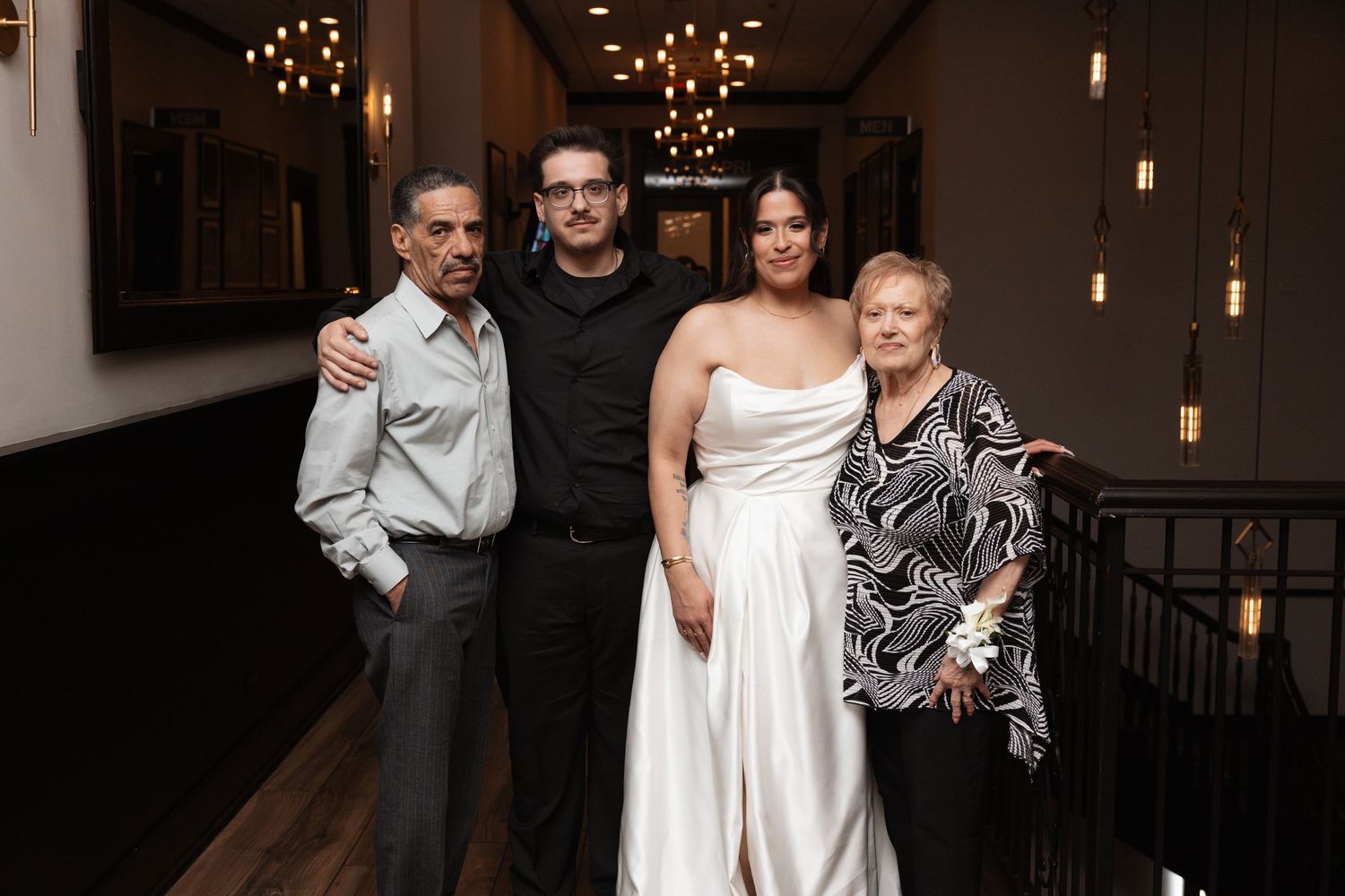 Bride poses with family members including parents on staircase inside warmly lit wedding reception venue.