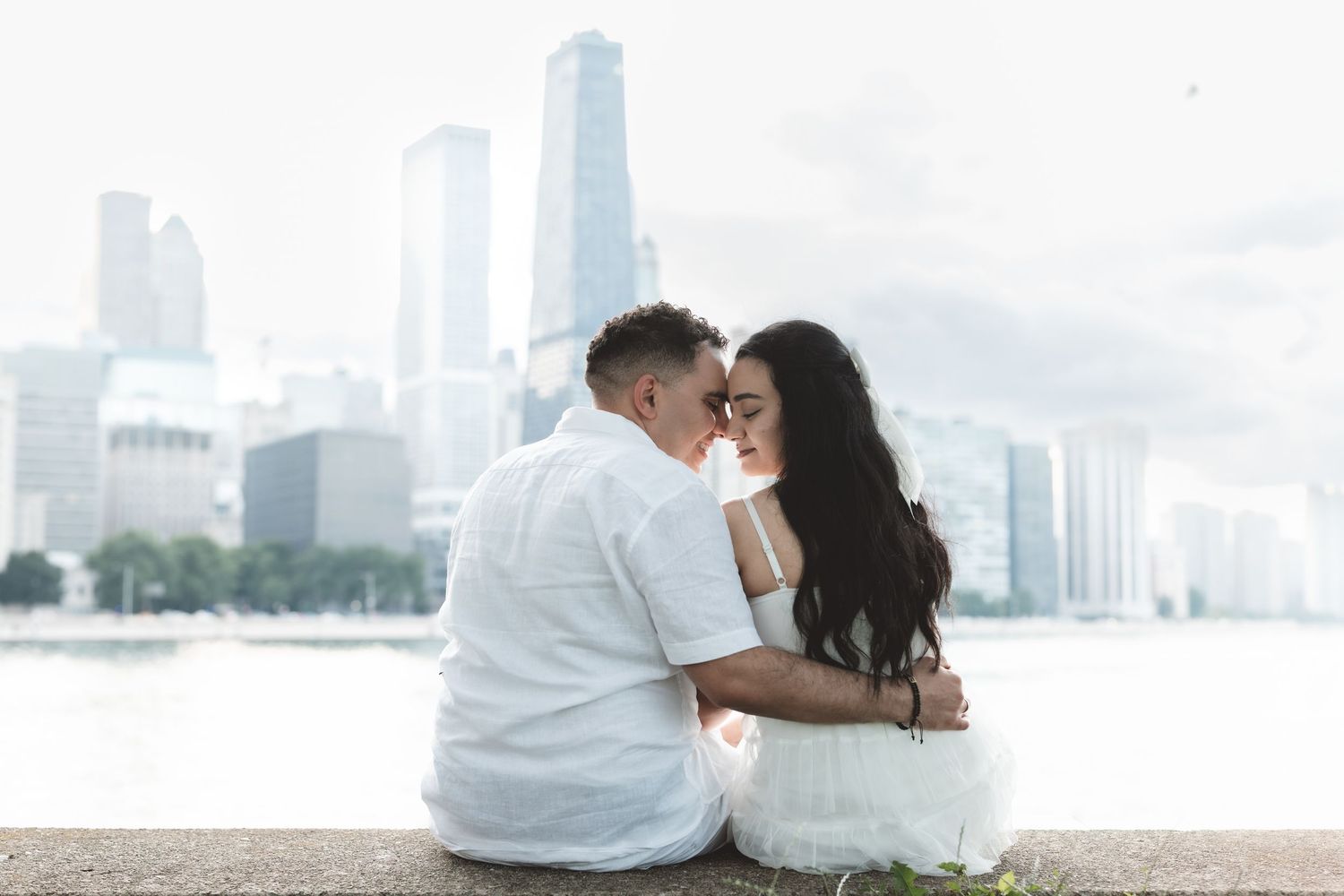 Romantic couple sitting close together by waterfront with city skyline in the background.