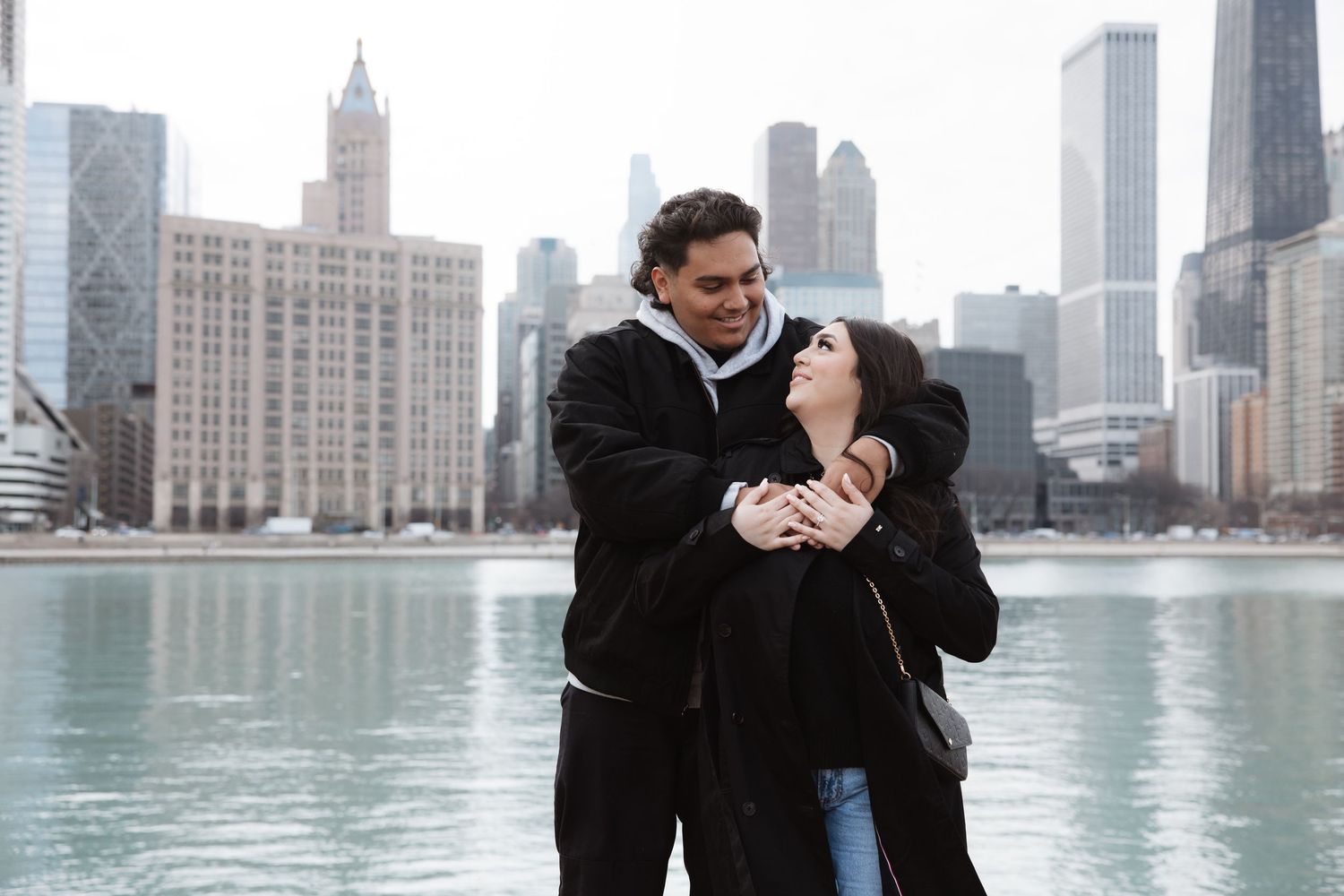 Happy couple hugging and smiling with the Chicago city skyline reflected in the lake behind them.
