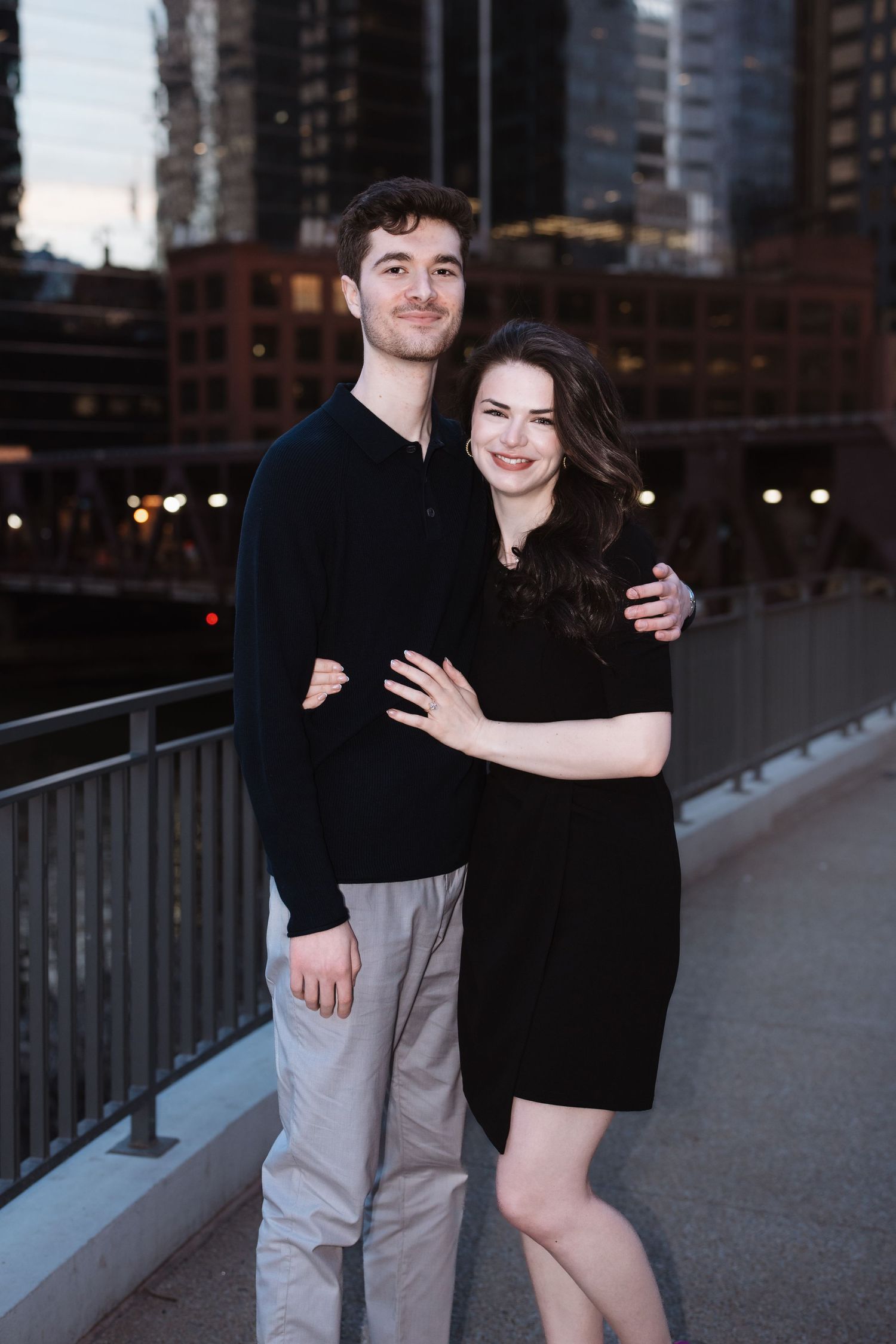 Smiling couple embracing on a bridge with Chicago city buildings lit up at dusk behind them.