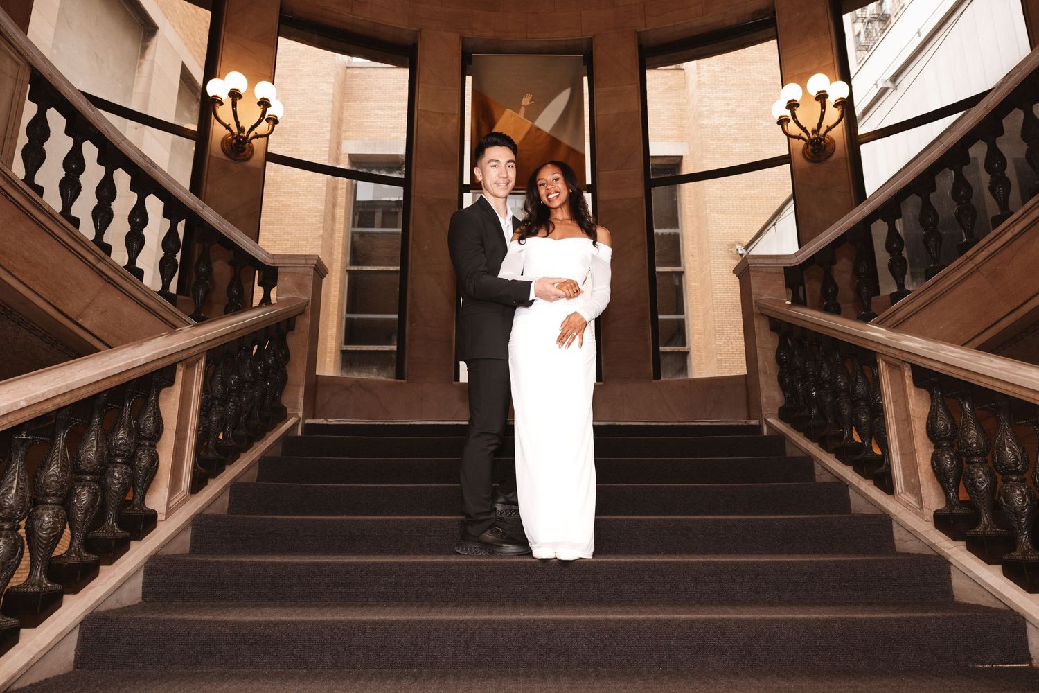Smiling newlyweds posing together on grand staircase flanked by warm wall sconces.
