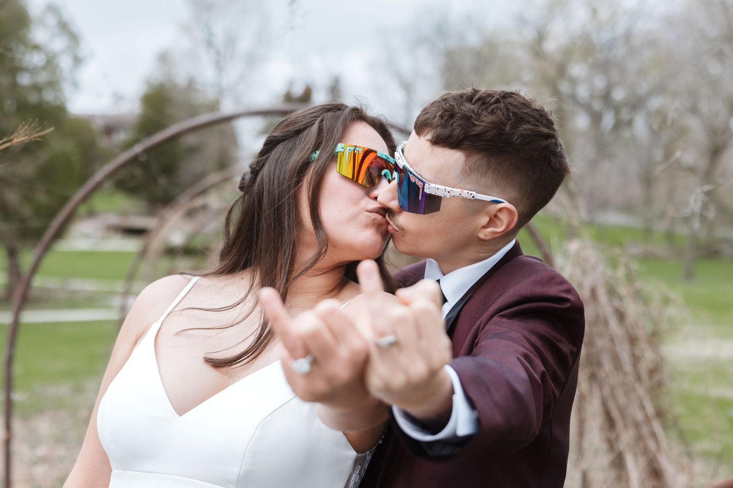 Newlywed couple kissing and showing wedding rings, wearing stylish sunglasses outdoors in a park setting.