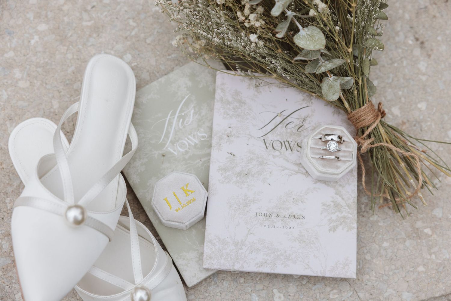 Elegant wedding flat lay with white bridal shoes, vow books, rings, and eucalyptus bouquet on marble surface.