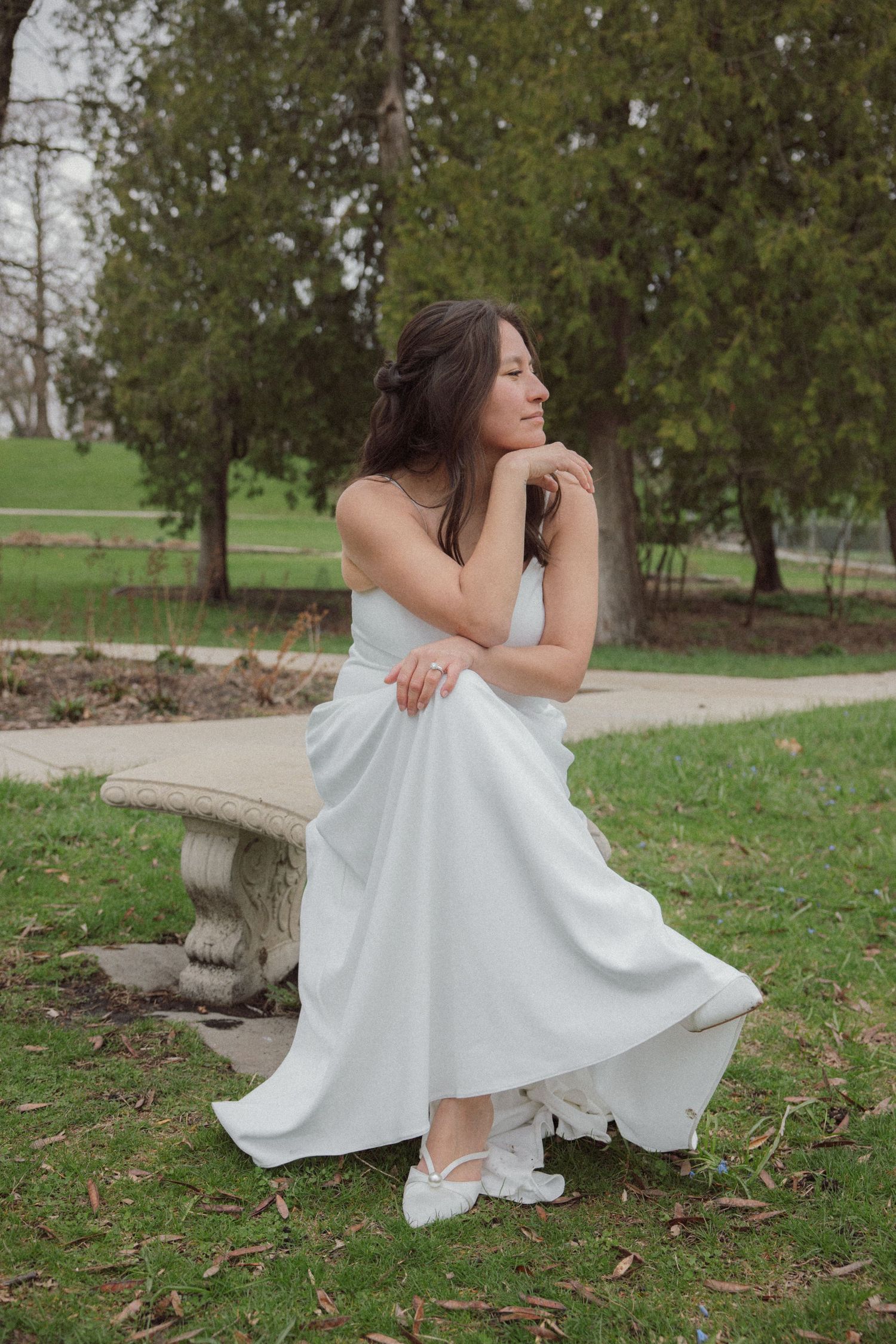 A bride in a white gown sits gracefully on a stone bench in a lush green park, gazing serenely into the distance.