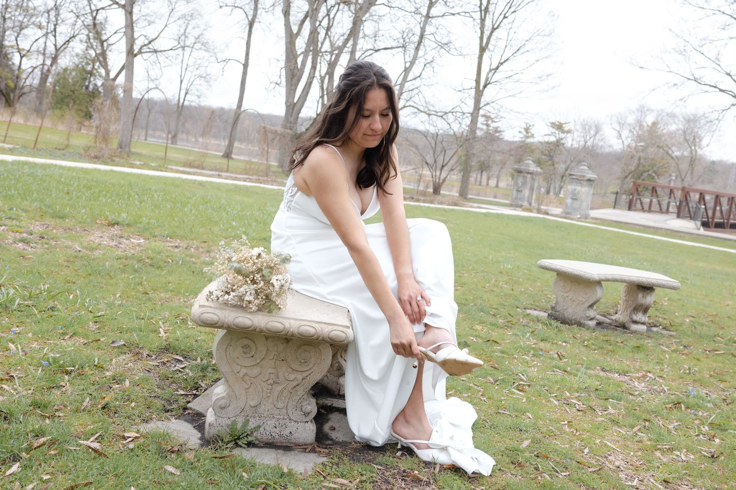 Bride in white gown sitting on stone bench in park, adjusting her shoe, with dried flowers beside her.