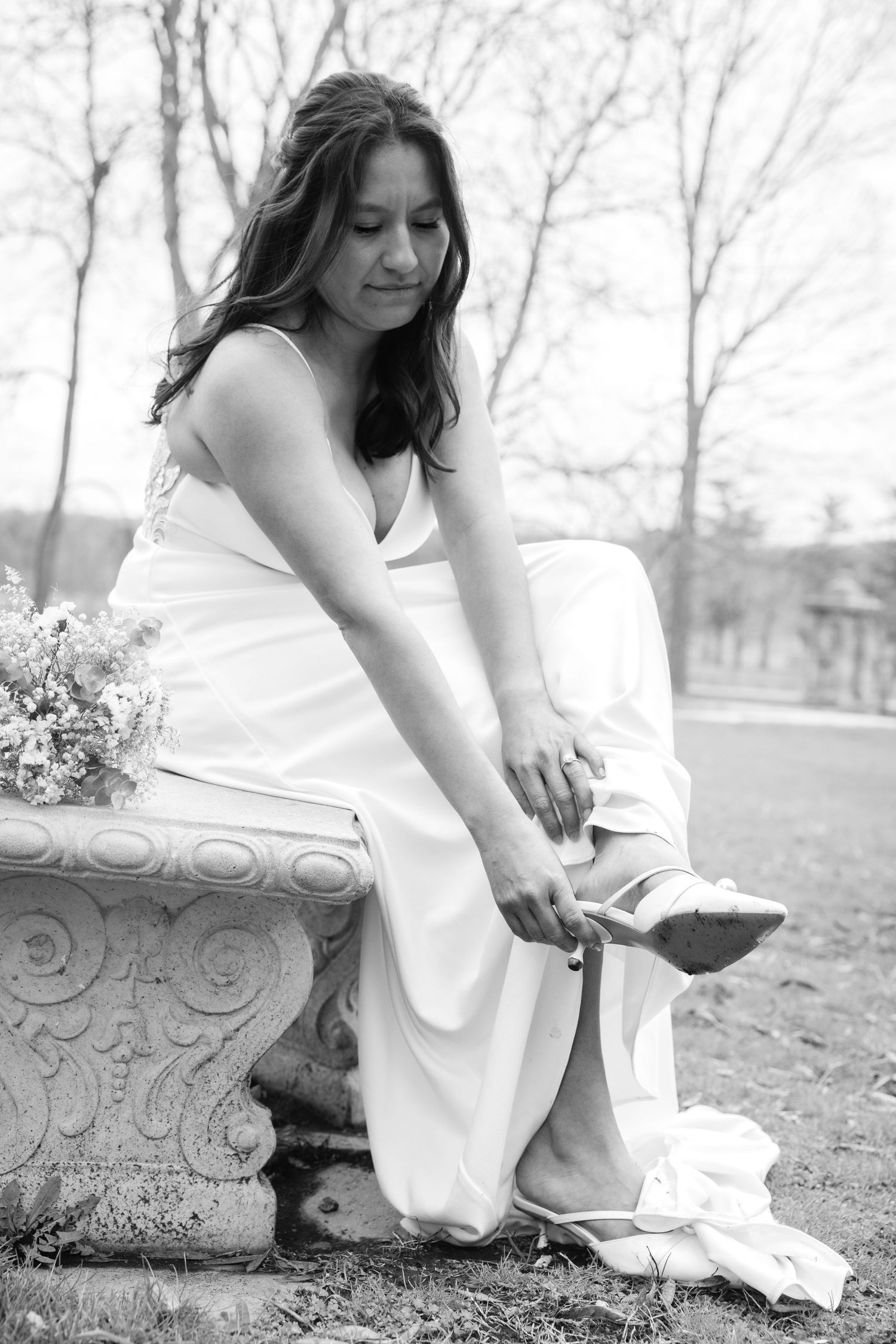 Bride in white dress sitting on ornate stone bench adjusting her heels outdoors, black and white wedding photo.