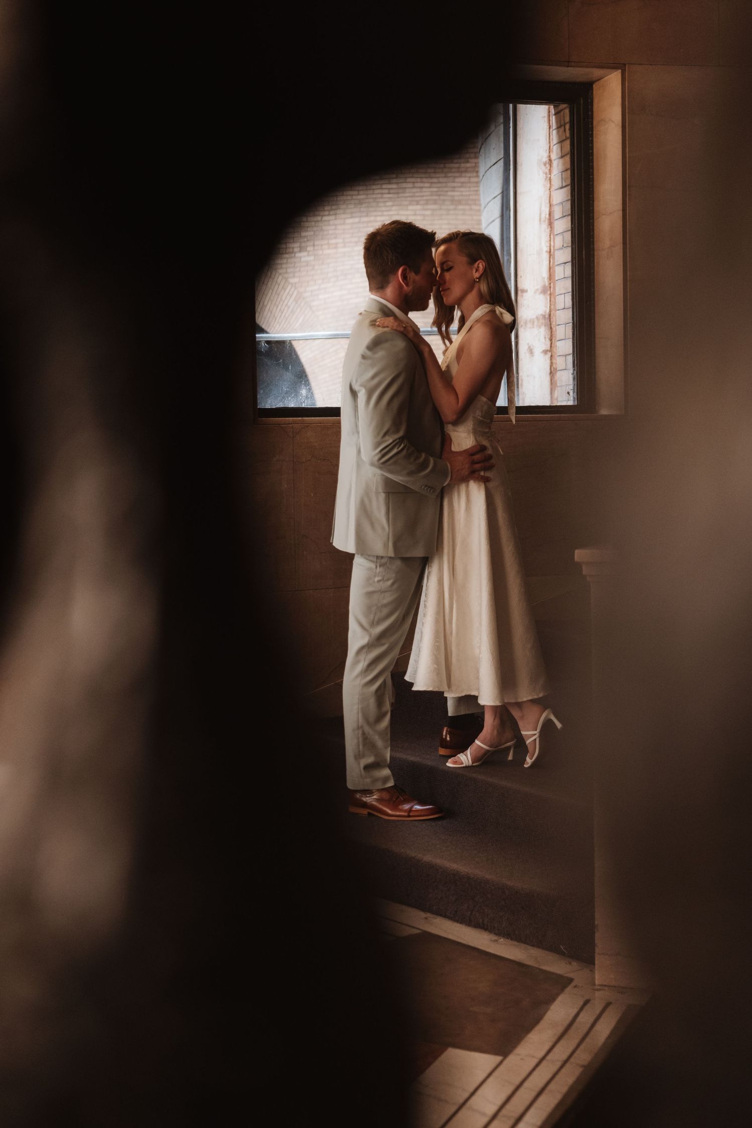 Newlyweds embracing near a window on a staircase landing, framed by dark architectural elements.