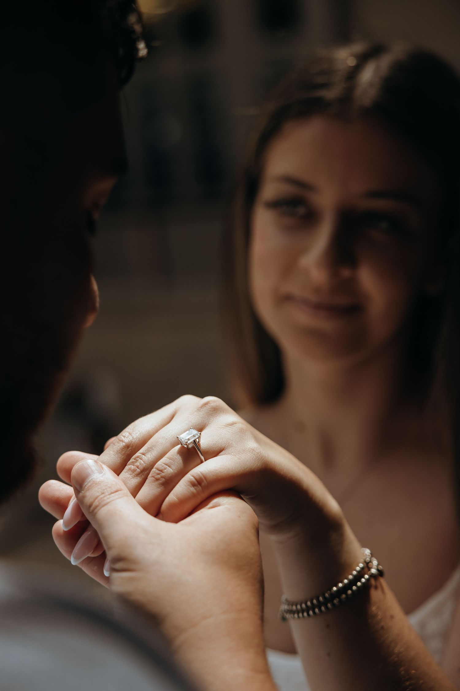 Engaged couple holding hands showing a sparkling diamond engagement ring, woman smiling softly in warm bokeh light.