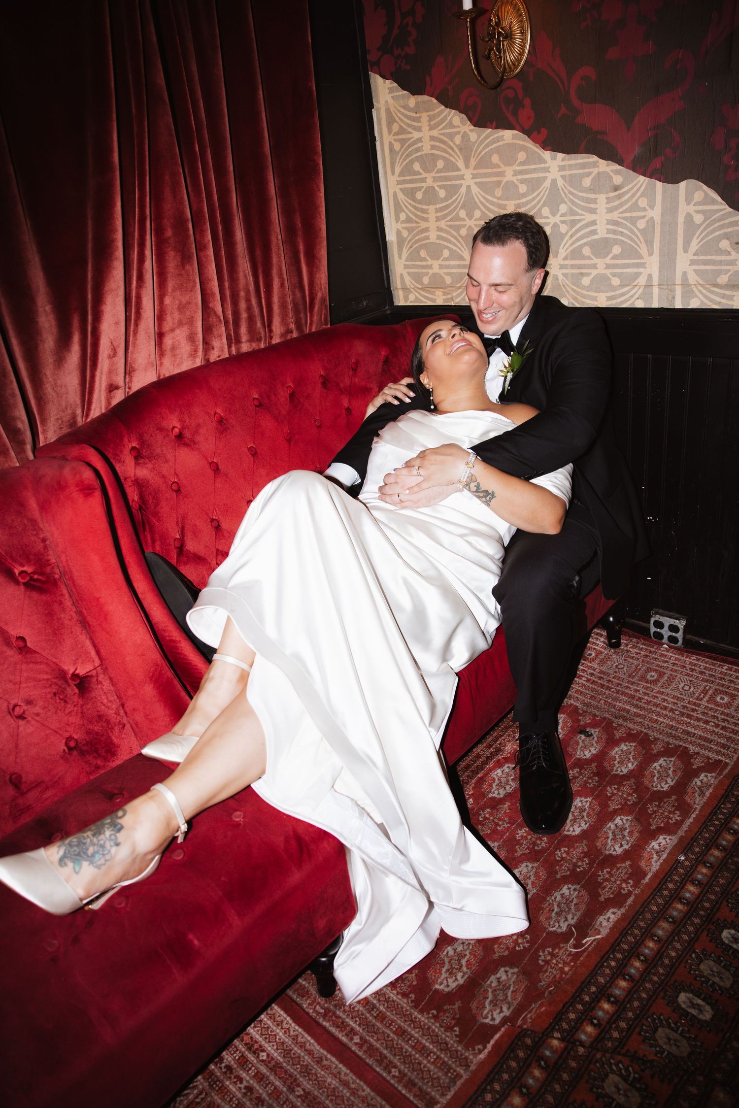 Bride reclines on a red velvet sofa as groom smiles and embraces her at wedding reception.