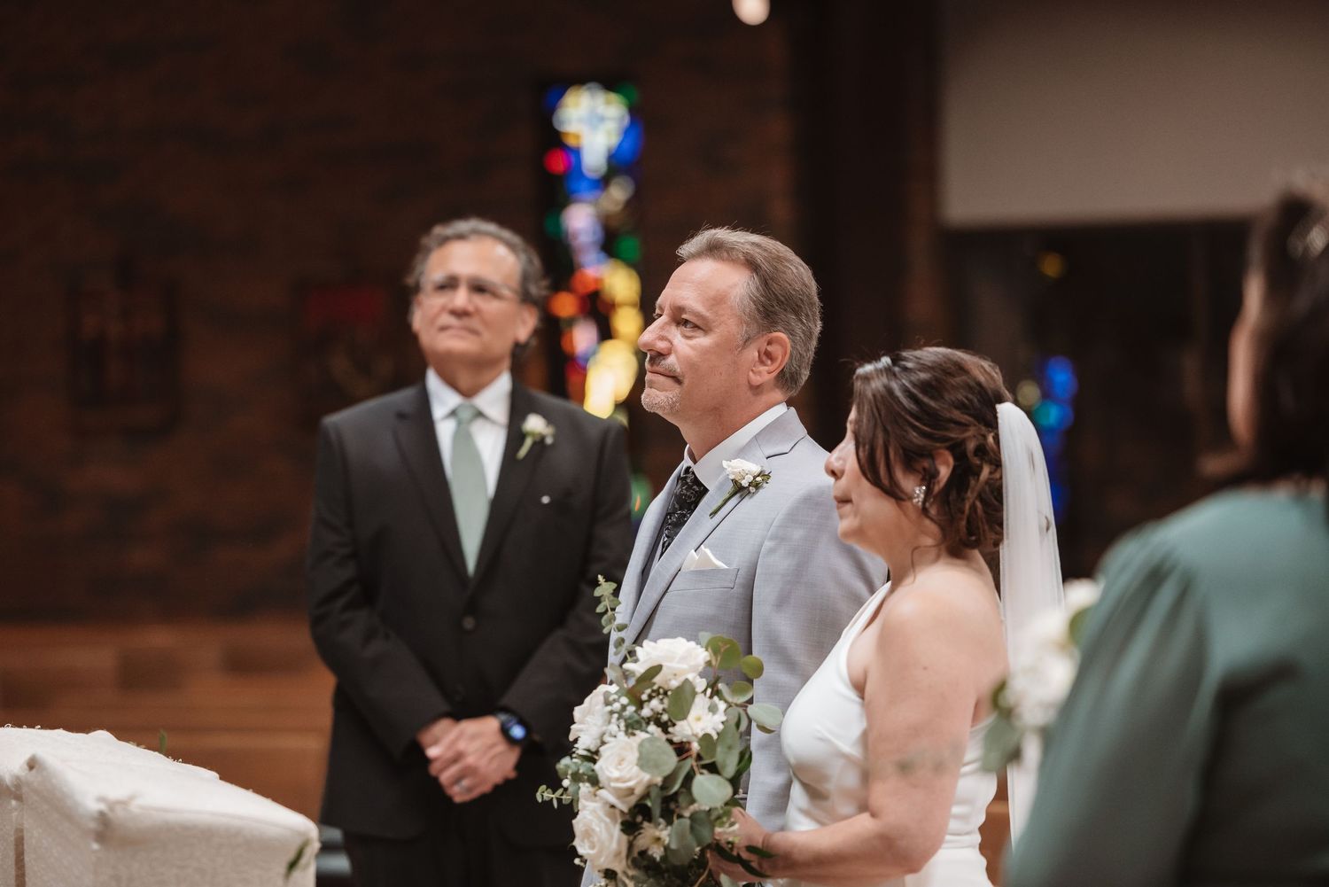 Bride and groom stand at church altar during wedding ceremony with stained glass windows in background.